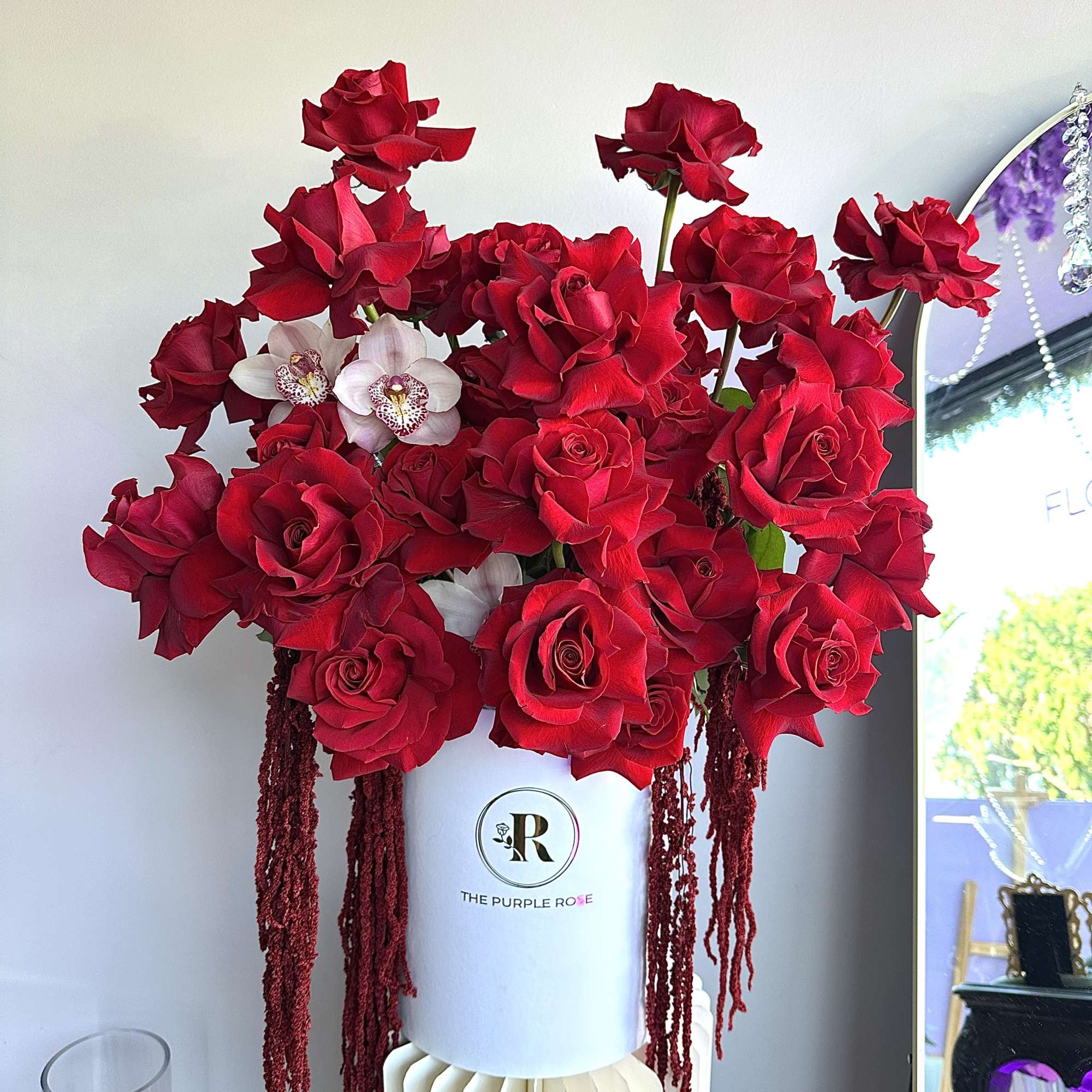 Tall arrangement of red roses with two pale orchids in a white hatbox-style container