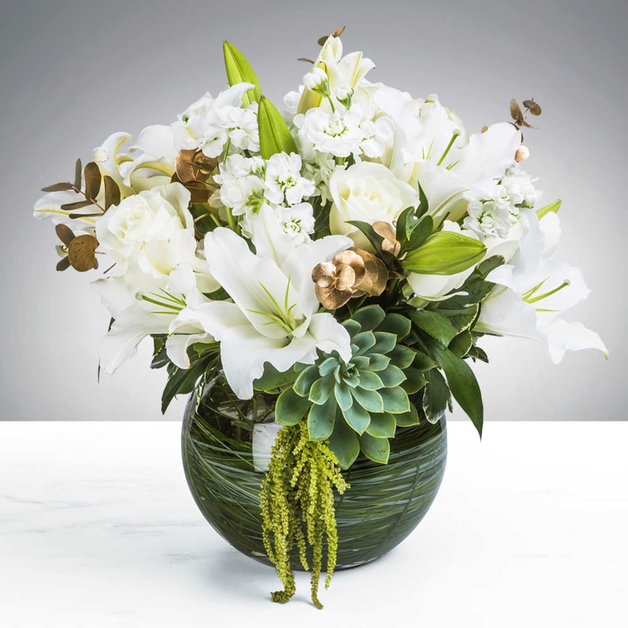 White lilies and roses arranged in a round glass vase with succulents