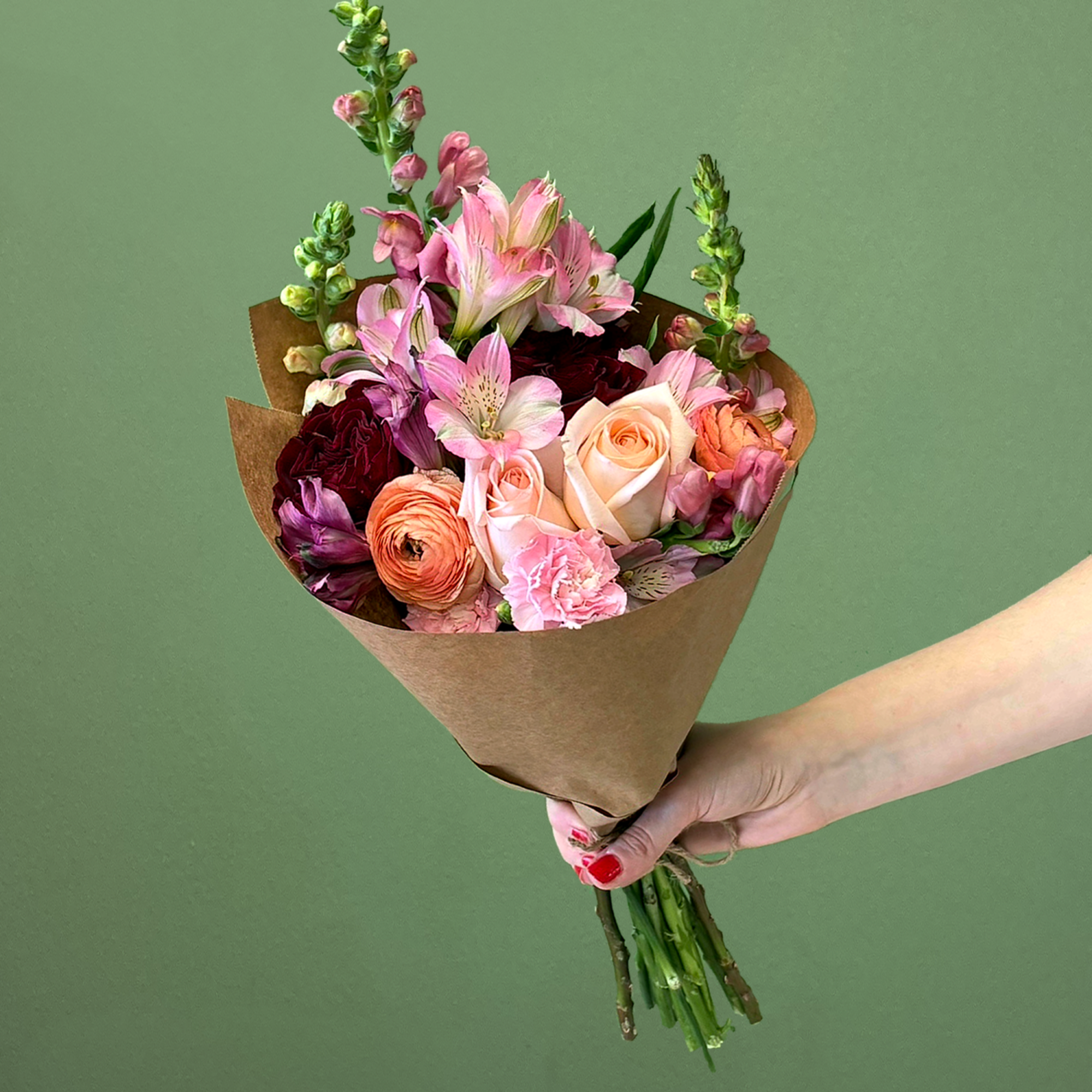 Hand-tied bouquet of pink and peach roses, ranunculus, and mixed blooms wrapped in brown paper.