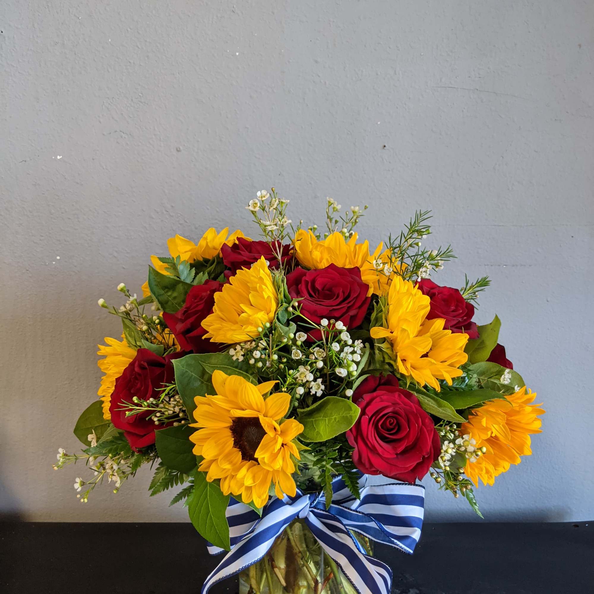 Arrangement of yellow sunflowers and red roses in a clear vase with a blue striped ribbon.