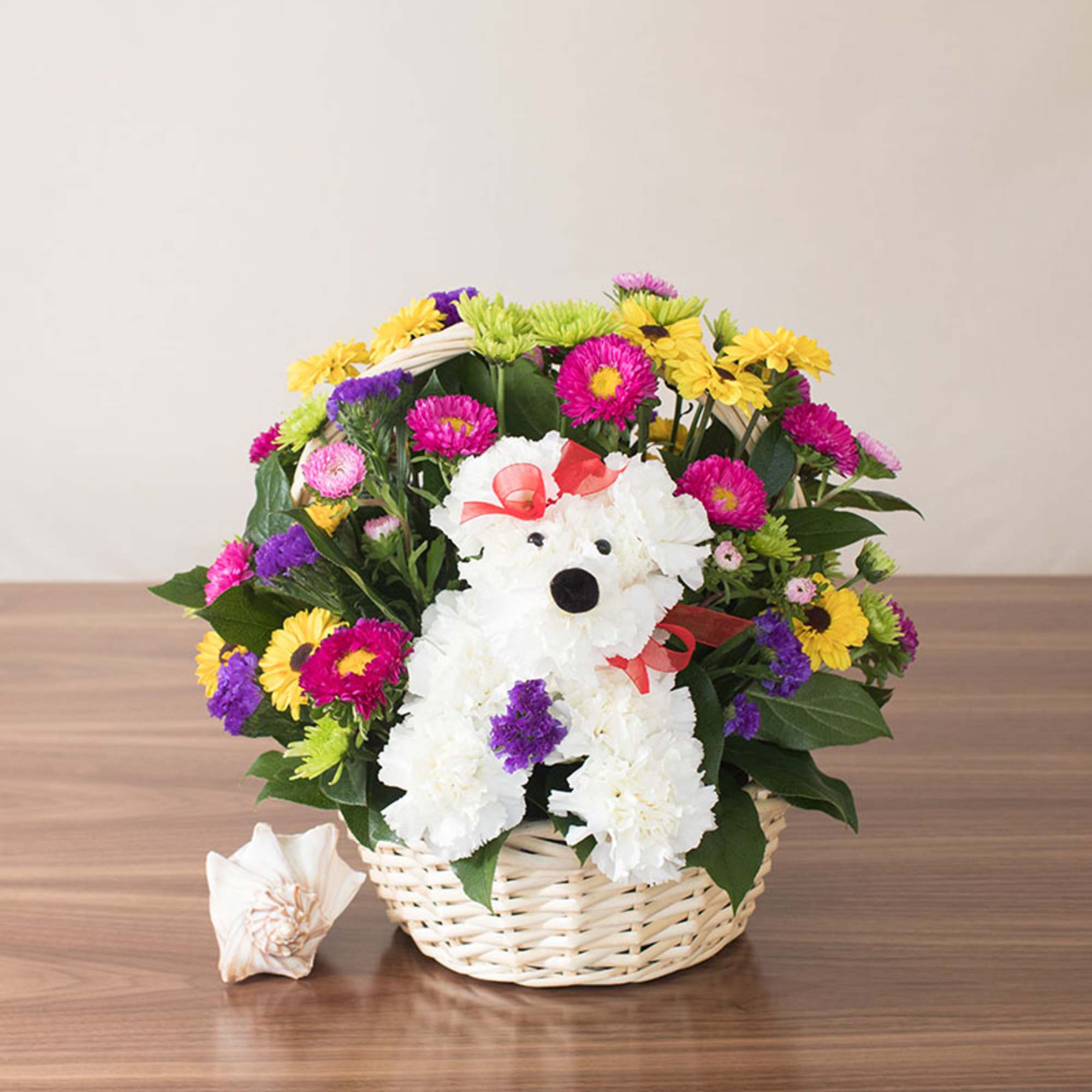 Basket arrangement of colorful daisies with a white carnation dog figure and red bow on a wooden table