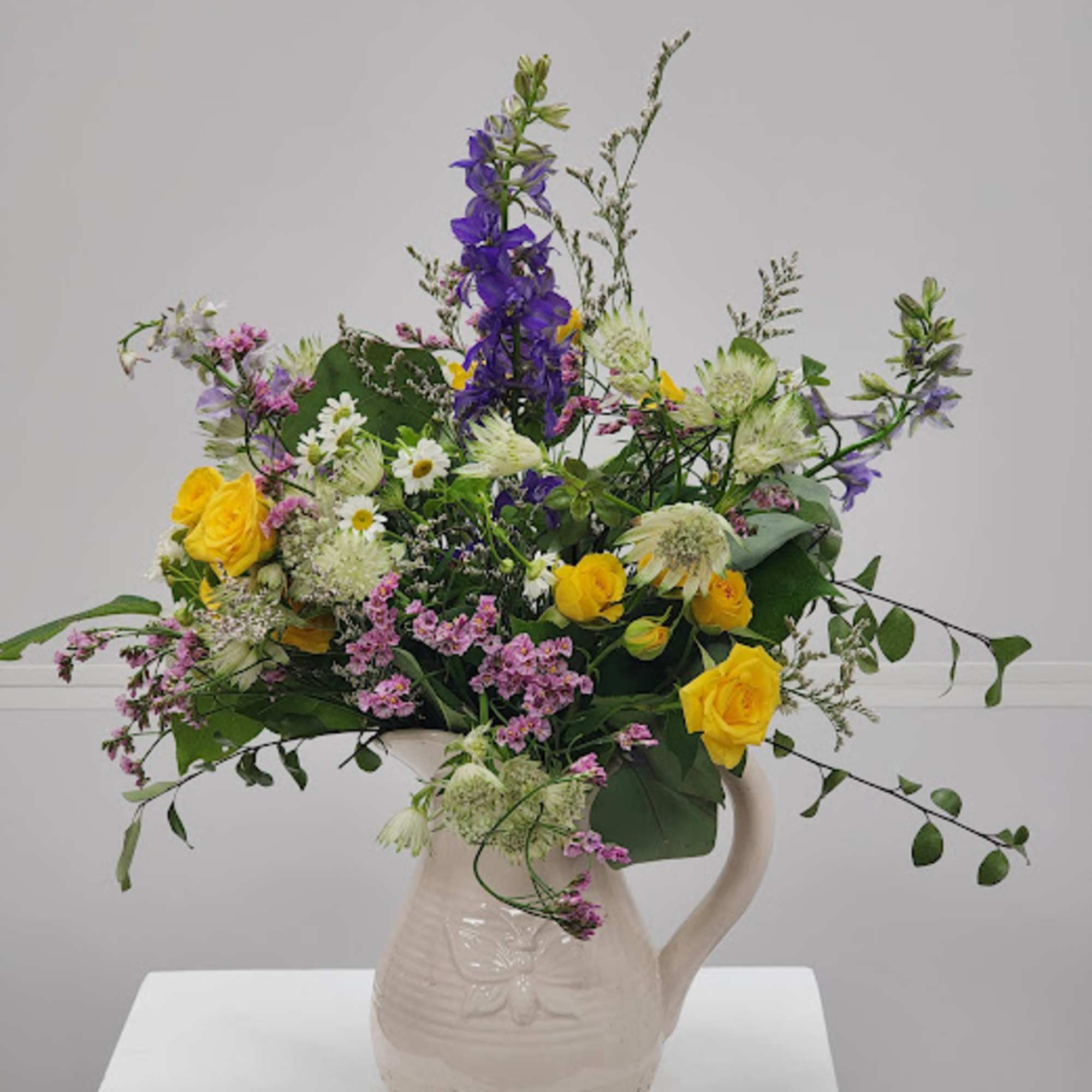 Mixed bouquet of yellow roses, purple blooms, and white flowers in a ceramic pitcher