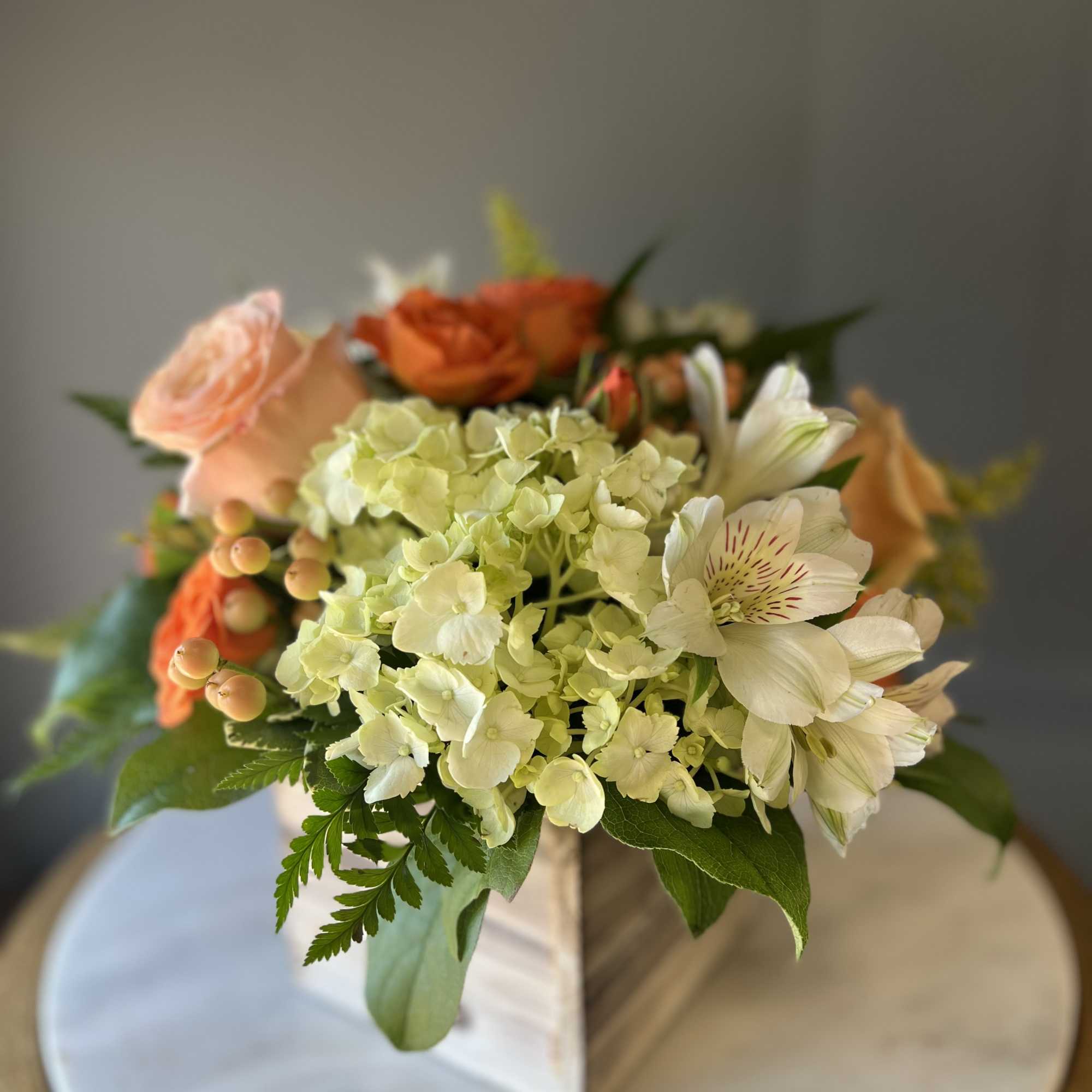 Low arrangement of green hydrangea, peach and orange roses, and white alstroemeria in a wooden cube vase