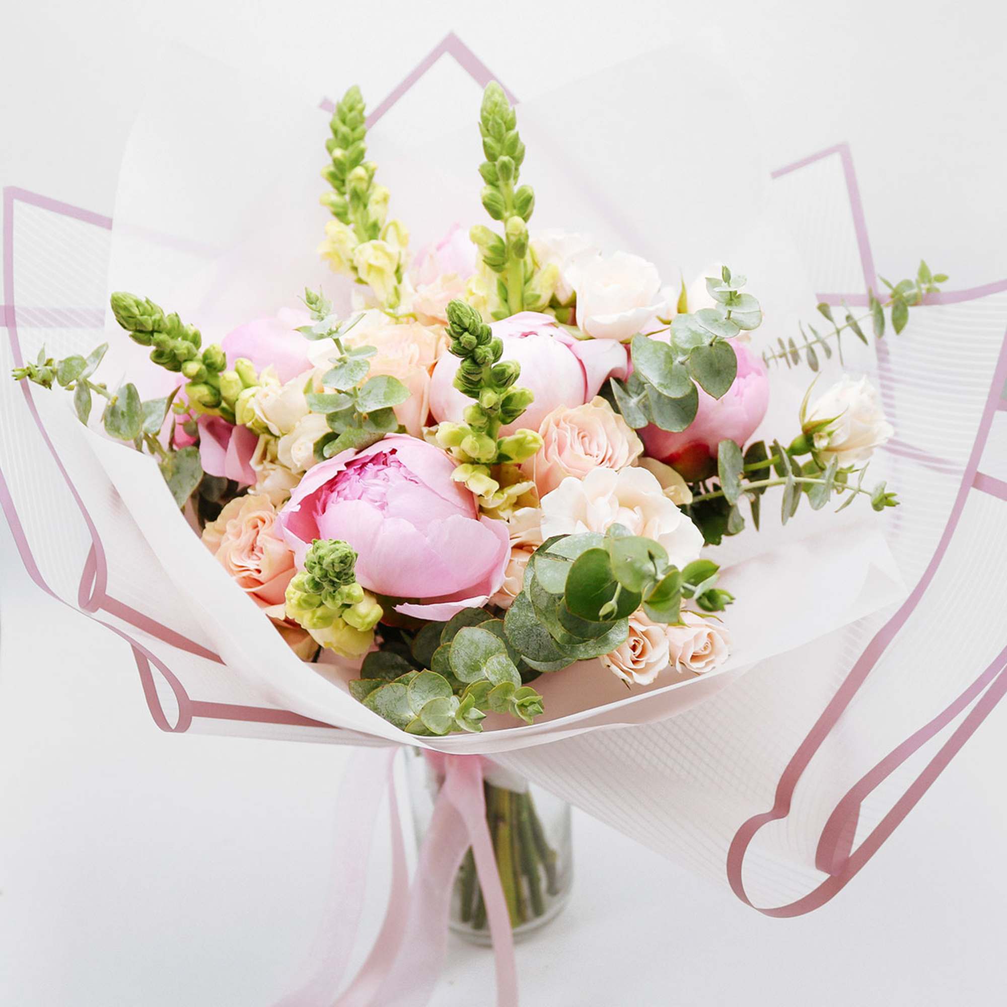Hand-tied bouquet of pink peonies, peach roses, and snapdragons wrapped in white paper in a glass vase