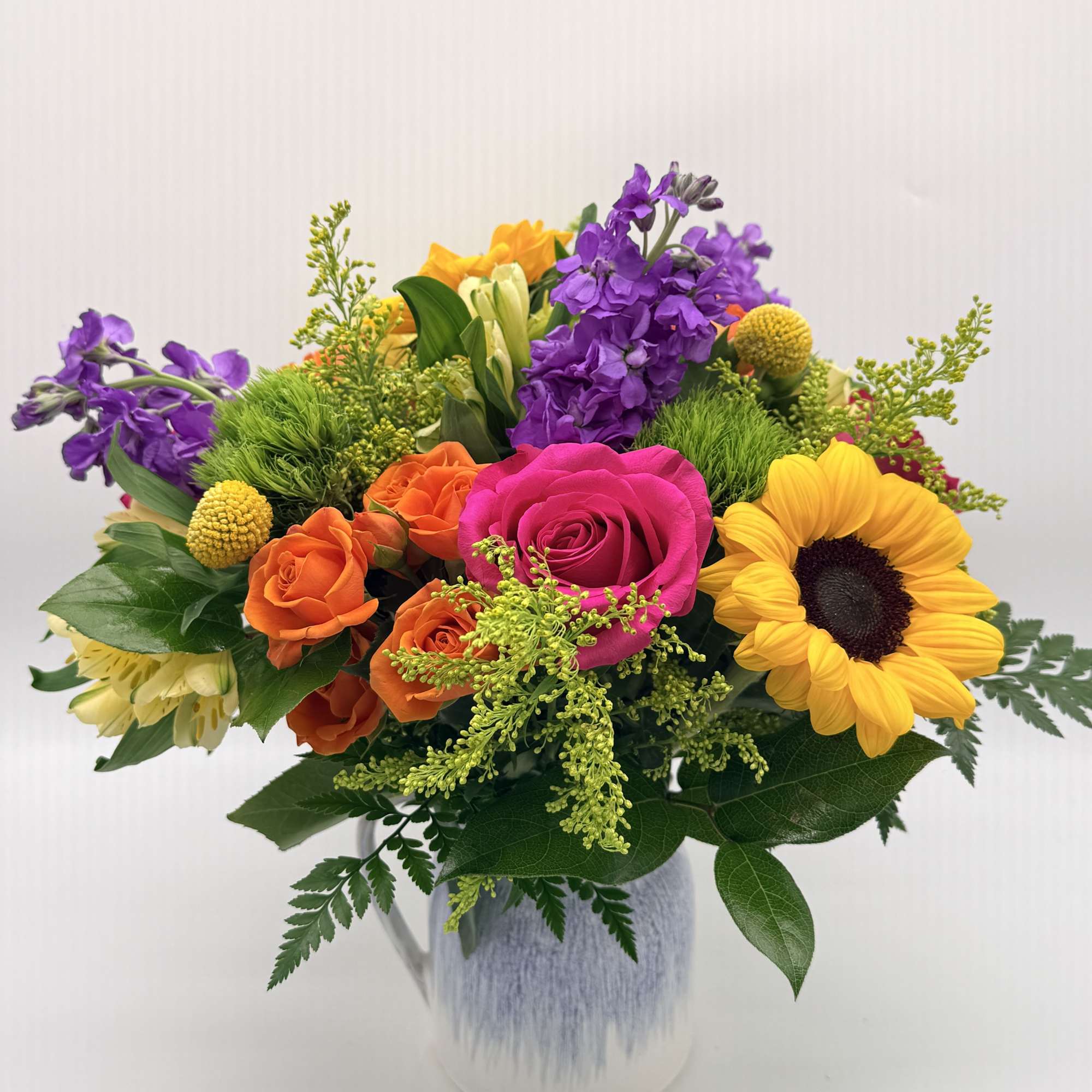 Bright mixed bouquet with sunflower, pink rose, orange and yellow blooms in a white ceramic pitcher.