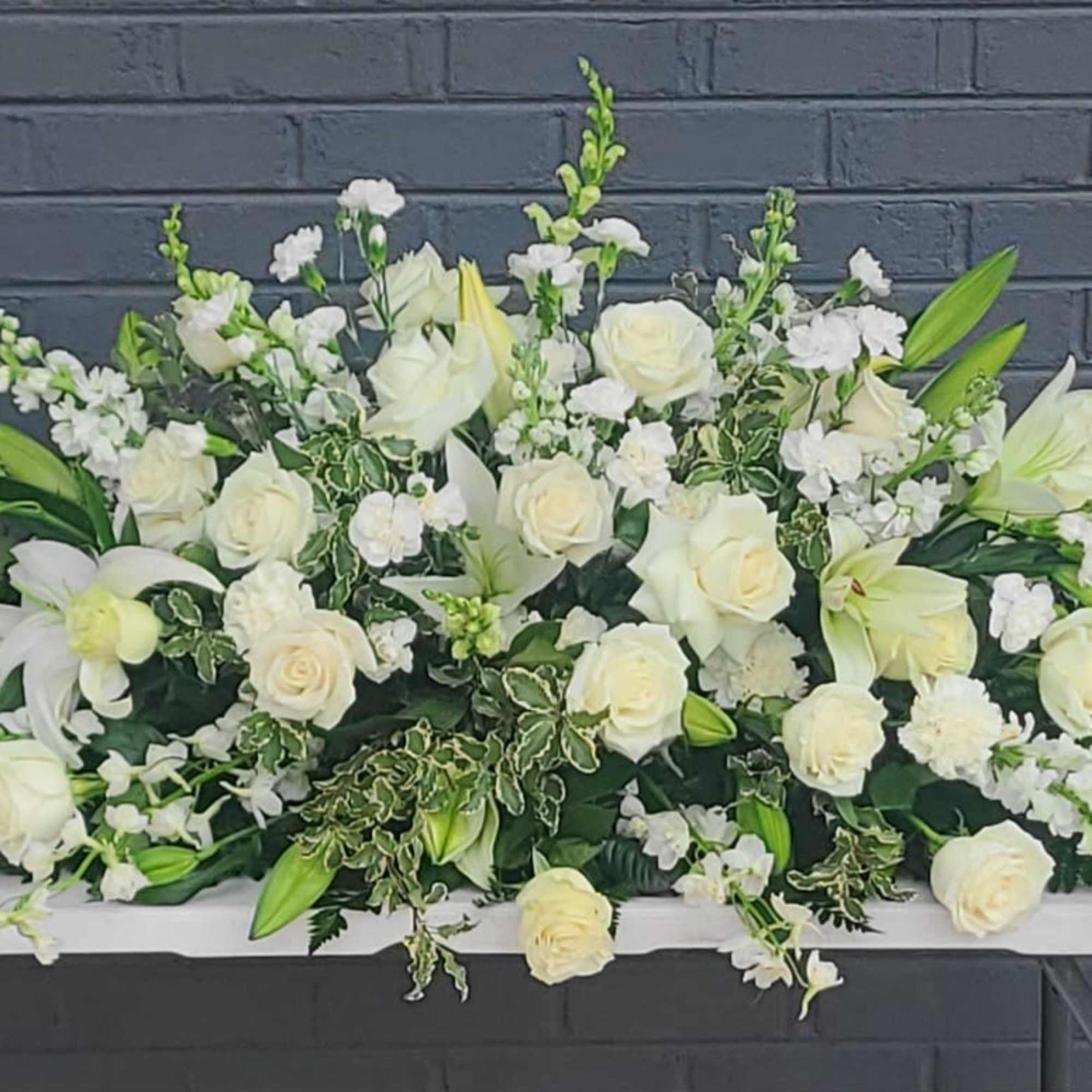 Cream-white roses and elegant white lilies form a serene, elongated casket spray