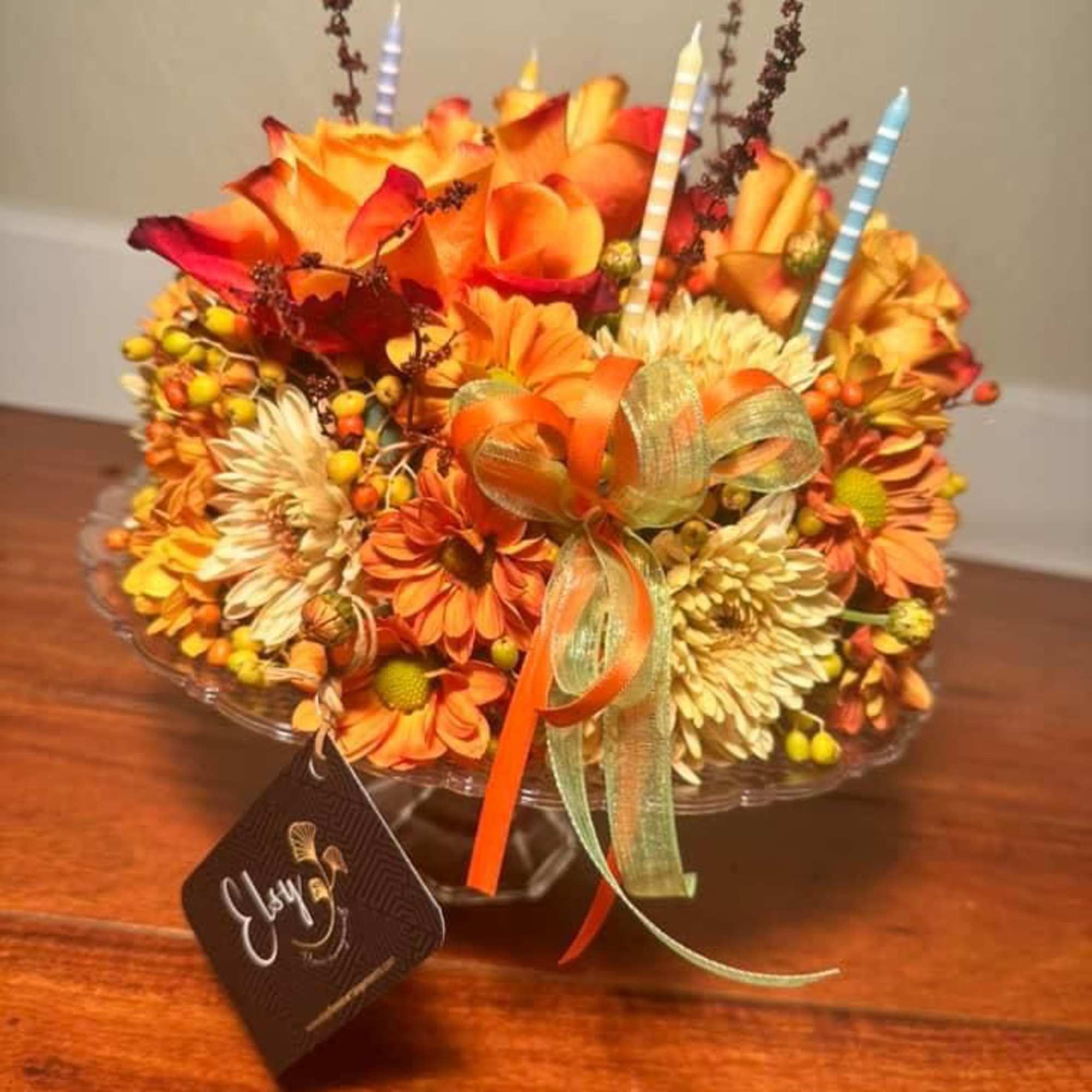 Orange and yellow floral arrangement with striped candles and a ribbon bow in a glass bowl