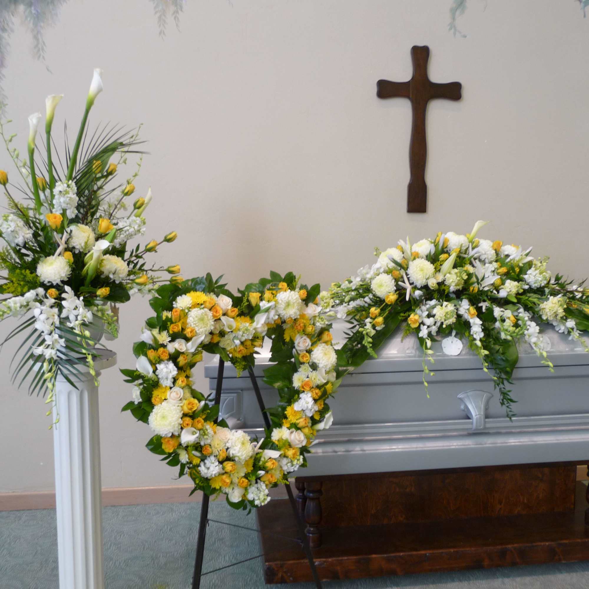 White and yellow funeral flowers arranged around a casket with a cross on the wall