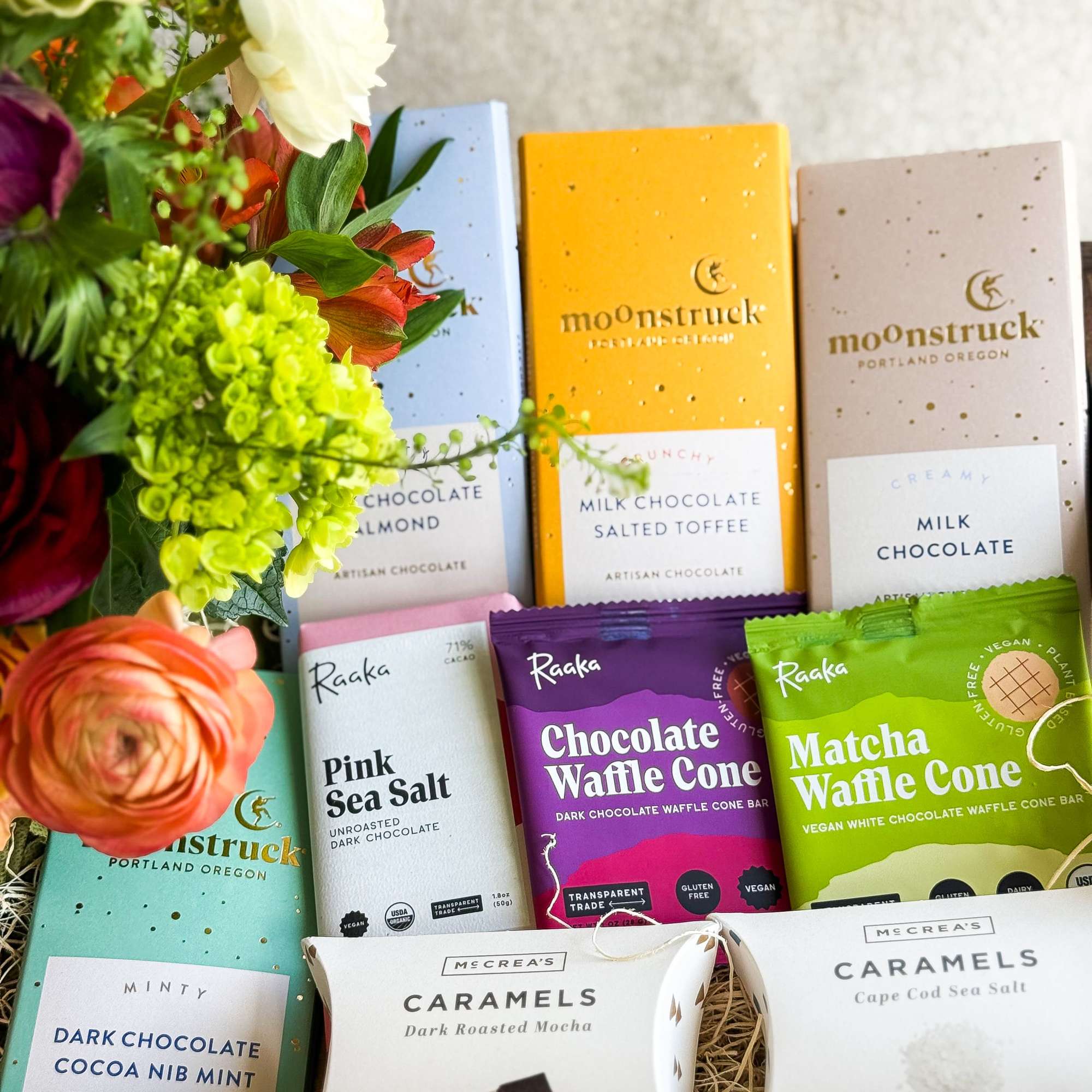 Flowers arranged beside assorted chocolate bars and caramels in a gift basket.
