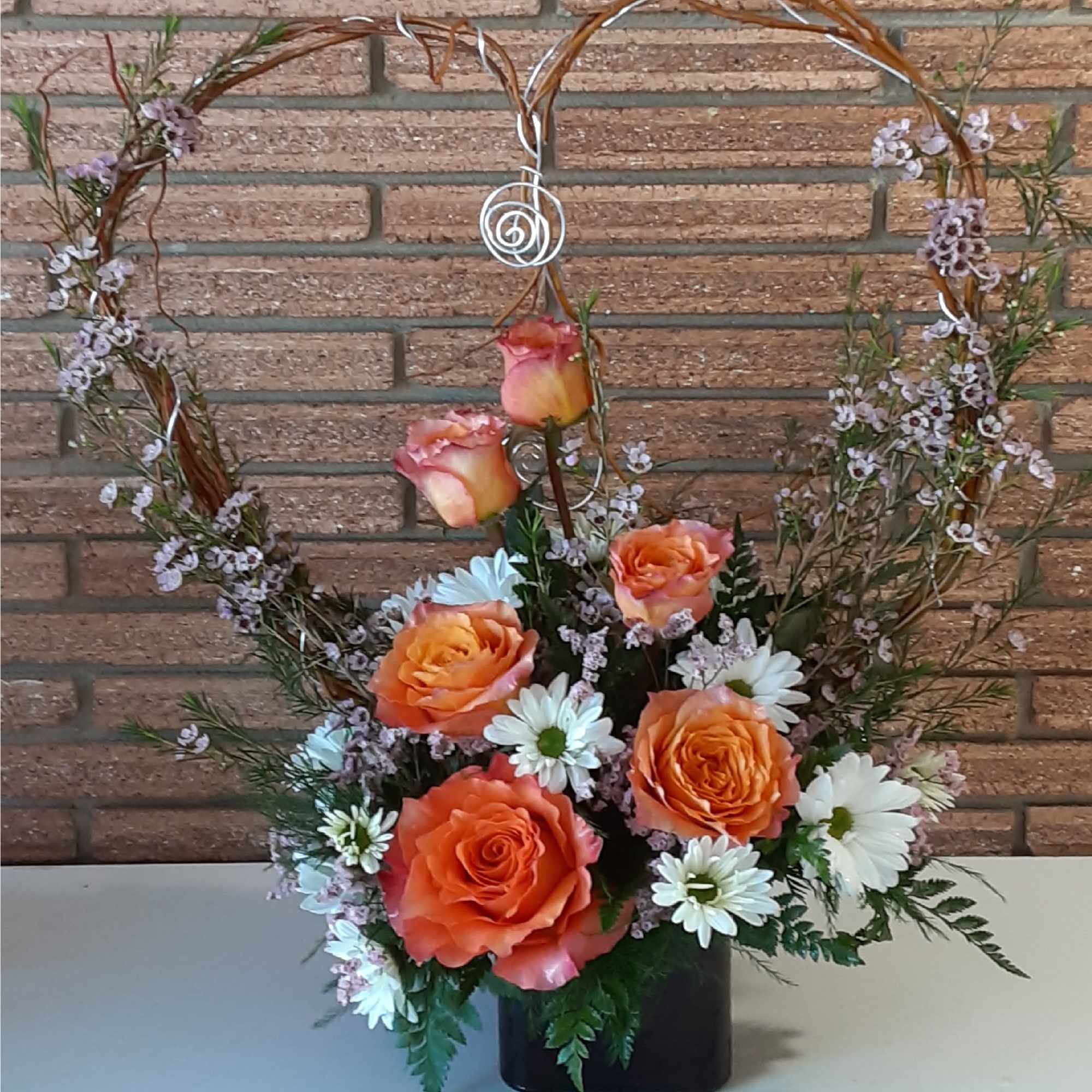 Orange roses and white daisies arranged in a black vase with a heart-shaped wire frame.