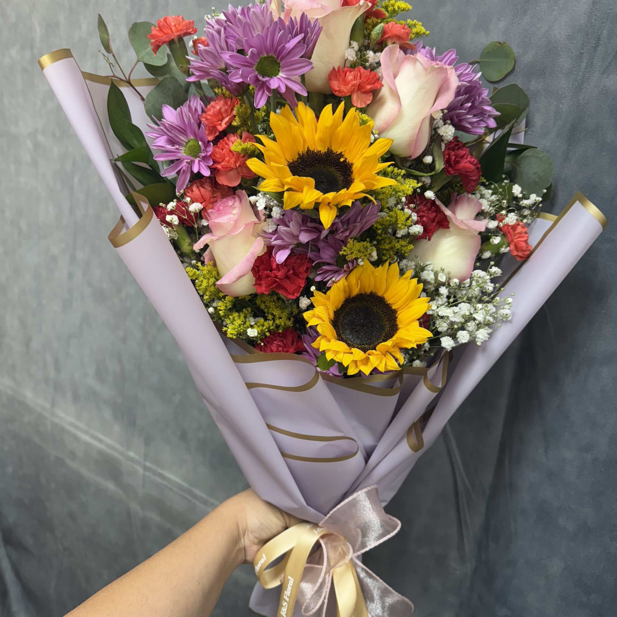 Bouquet of sunflowers, pink roses, and purple daisies wrapped in lavender paper