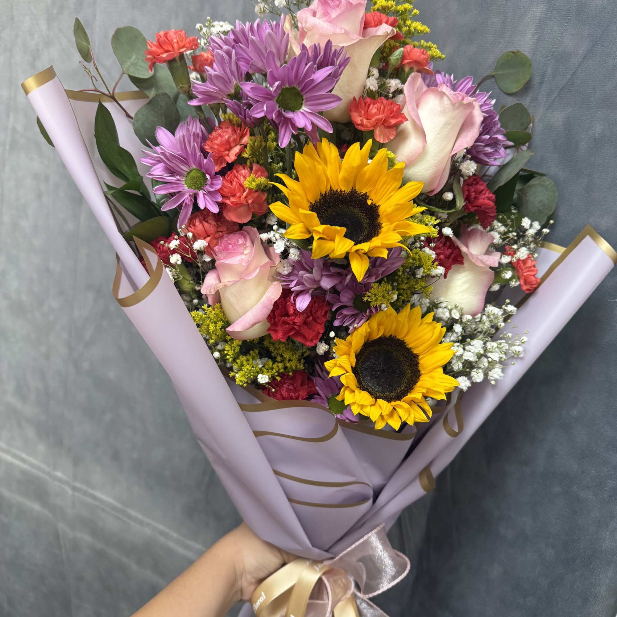Handheld bouquet of sunflowers, pink roses, and purple daisies wrapped in lavender paper