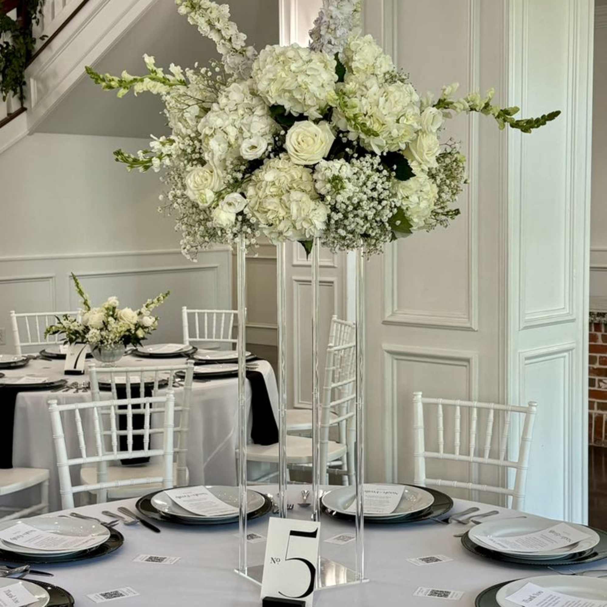 Tall white floral centerpiece on a clear stand above a set dining table