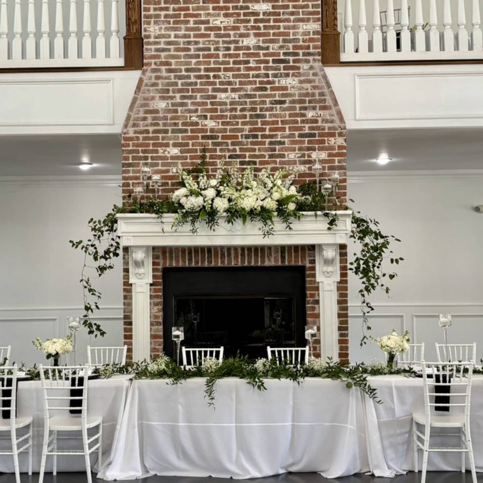 White floral arrangements decorate a fireplace mantel and long table in a reception hall.