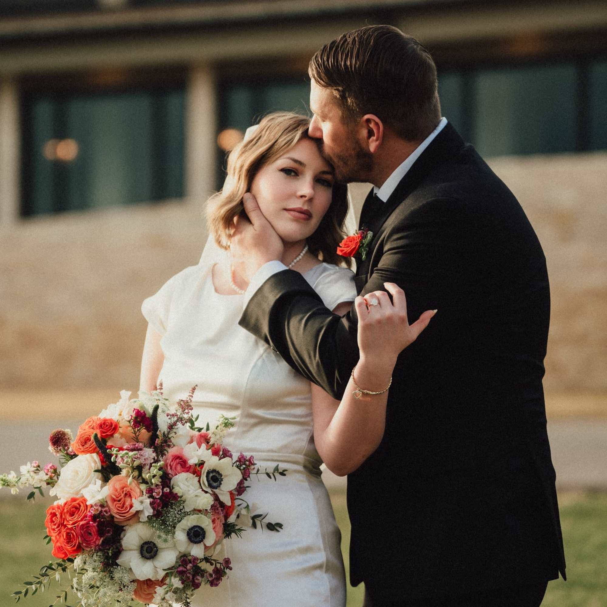 Bride holding a colorful bouquet while a groom kisses her forehead