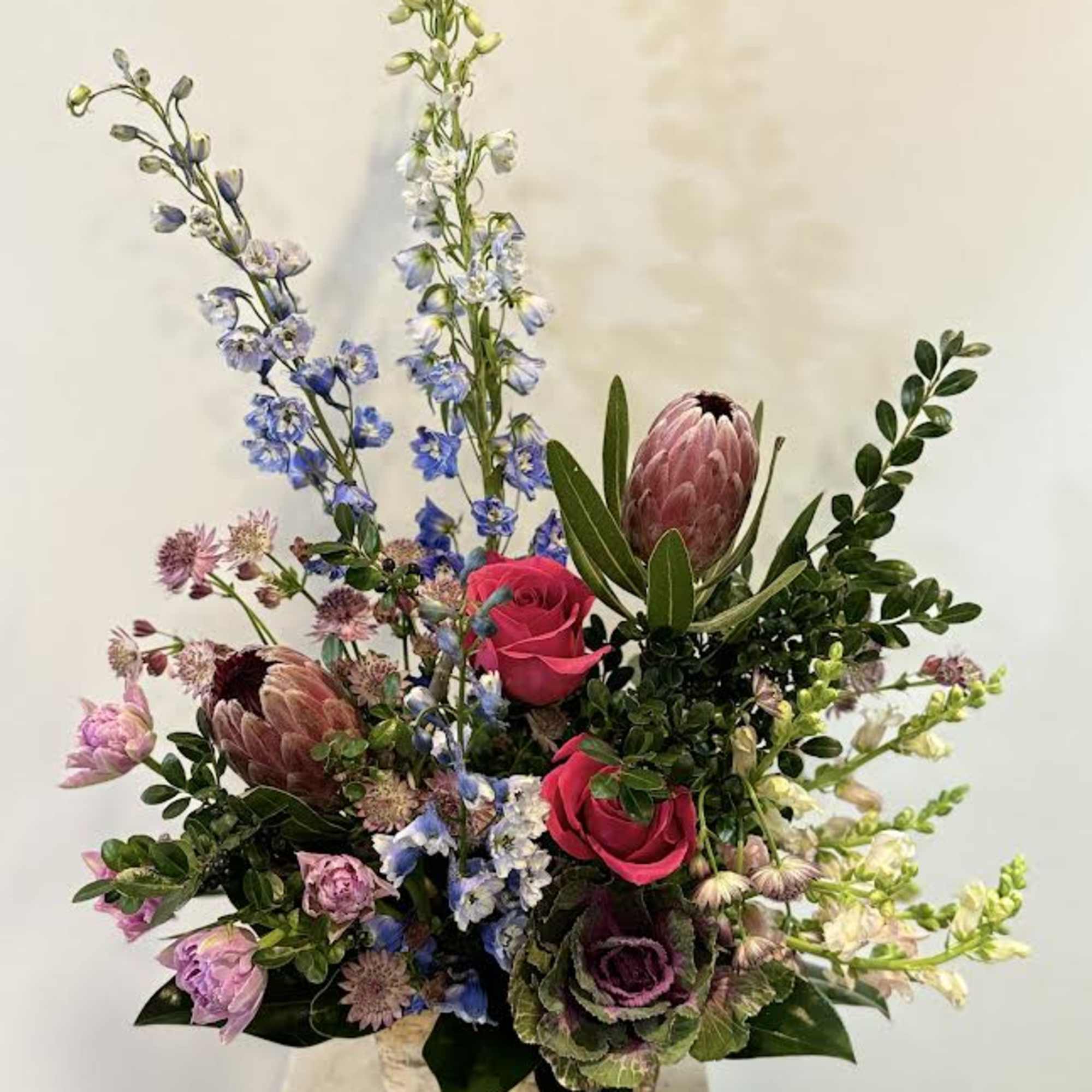 Tall mixed bouquet with pink roses, blue delphinium, and protea in a square vase