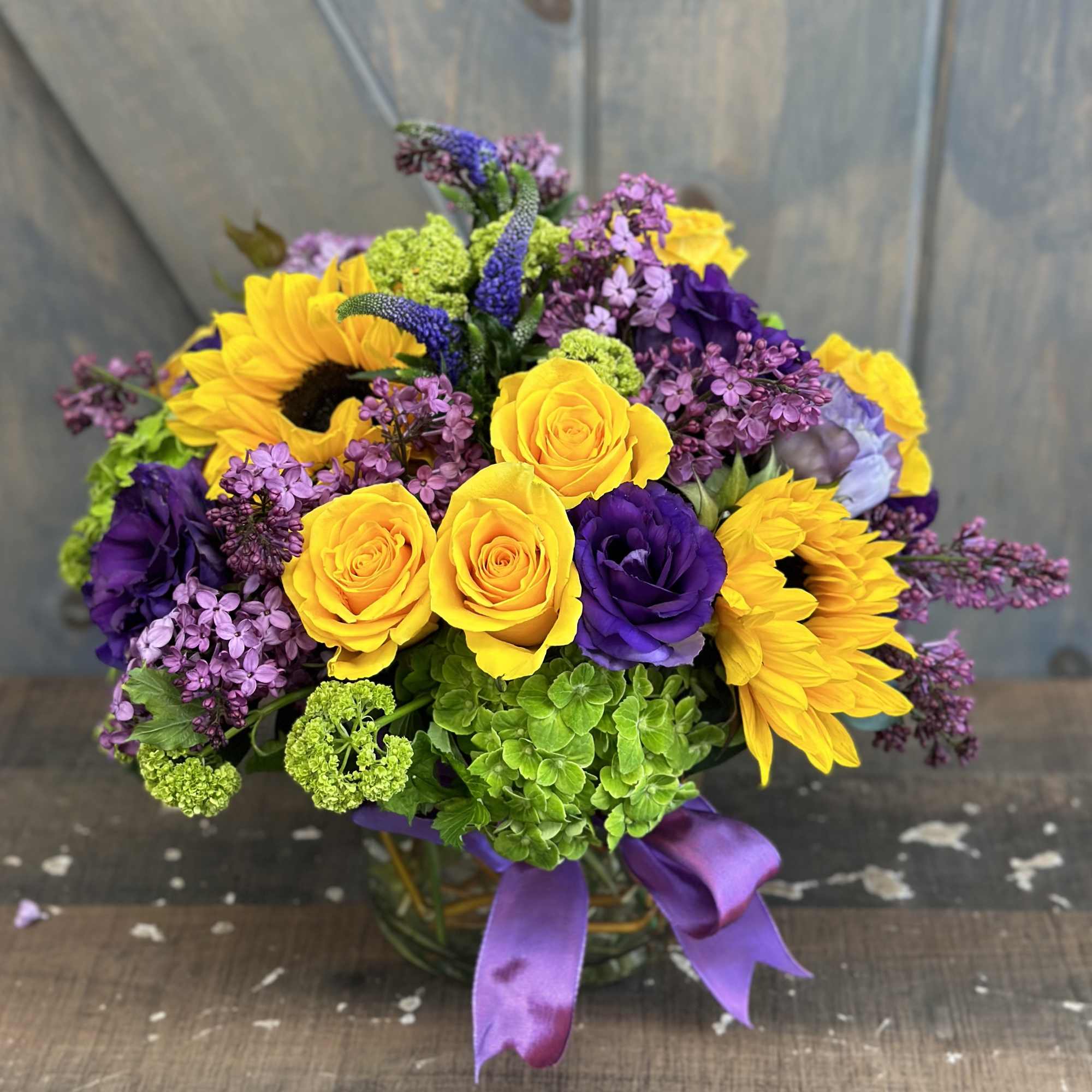 Bouquet of yellow roses, sunflowers, and purple flowers in a glass vase