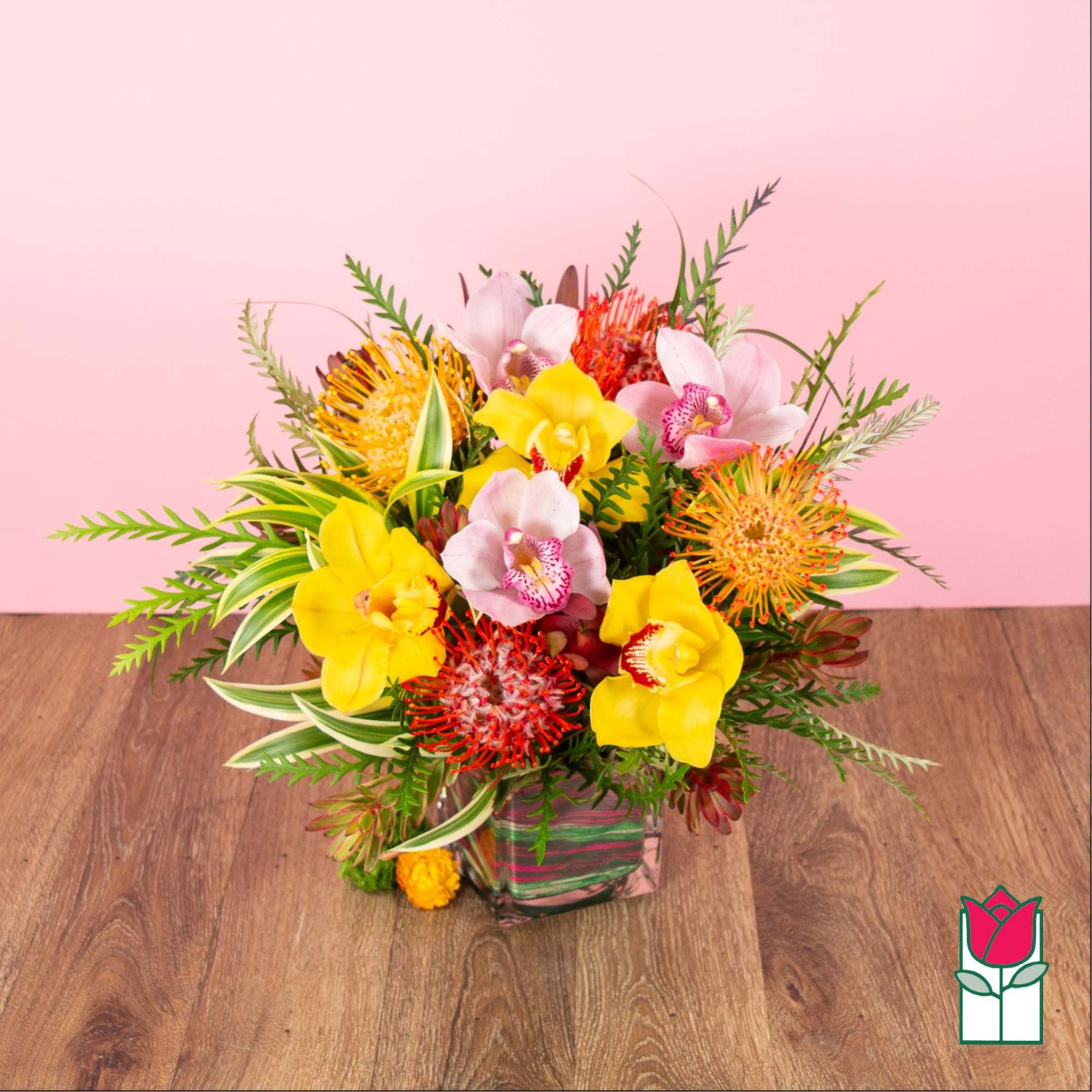 Bright arrangement of yellow and pink orchids with orange protea in a glass cube vase