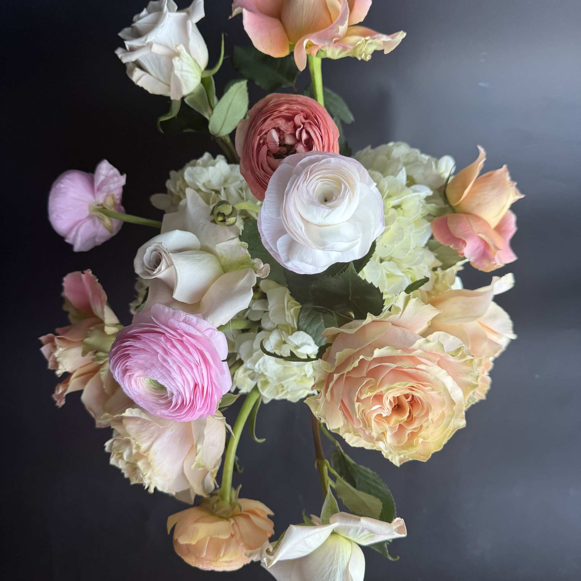 Arrangement of peach roses, white hydrangeas, and pink ranunculus in a compact design on a dark background