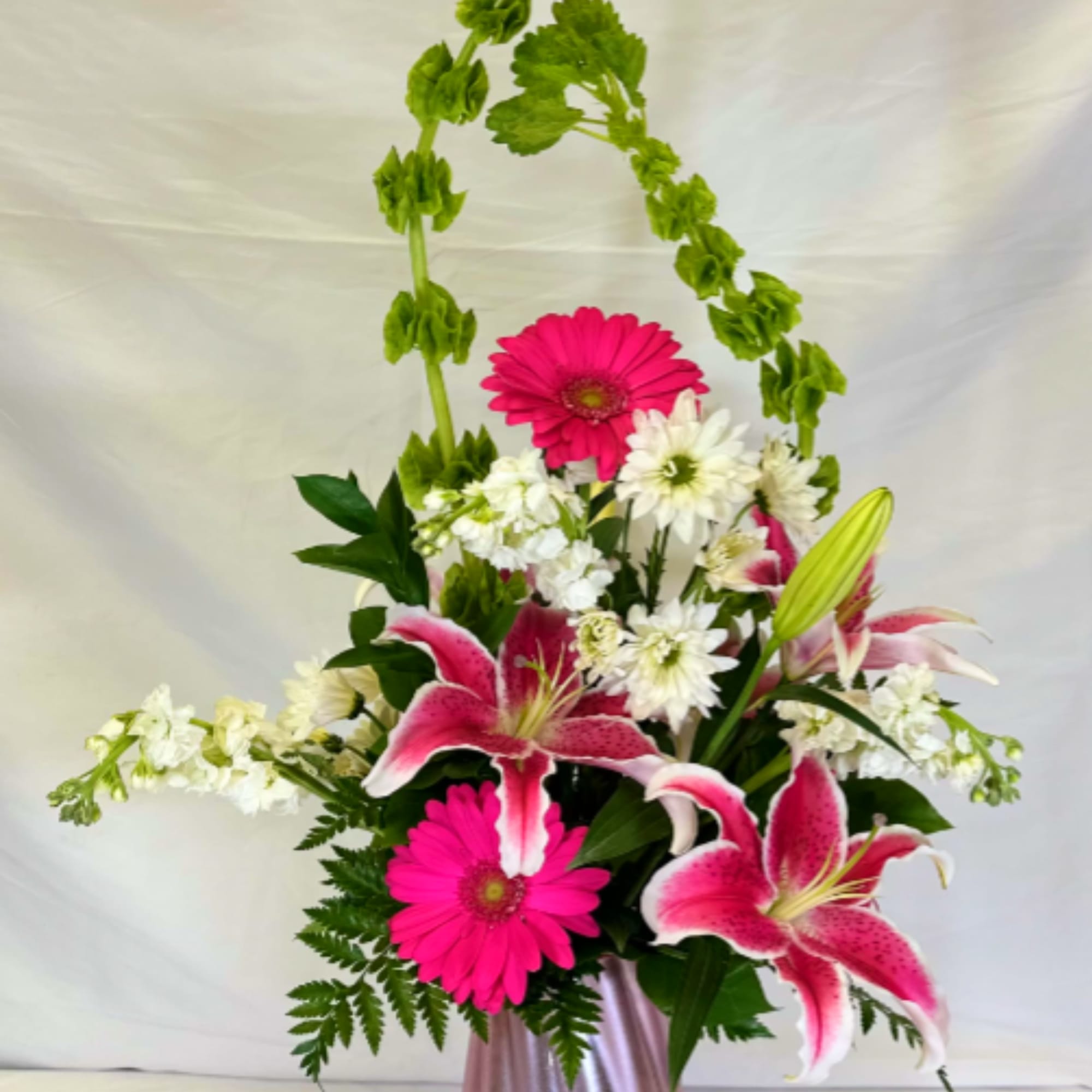 Tall pink and white arrangement with lilies and gerbera daisies in a pink vase with arching green bells of Ireland