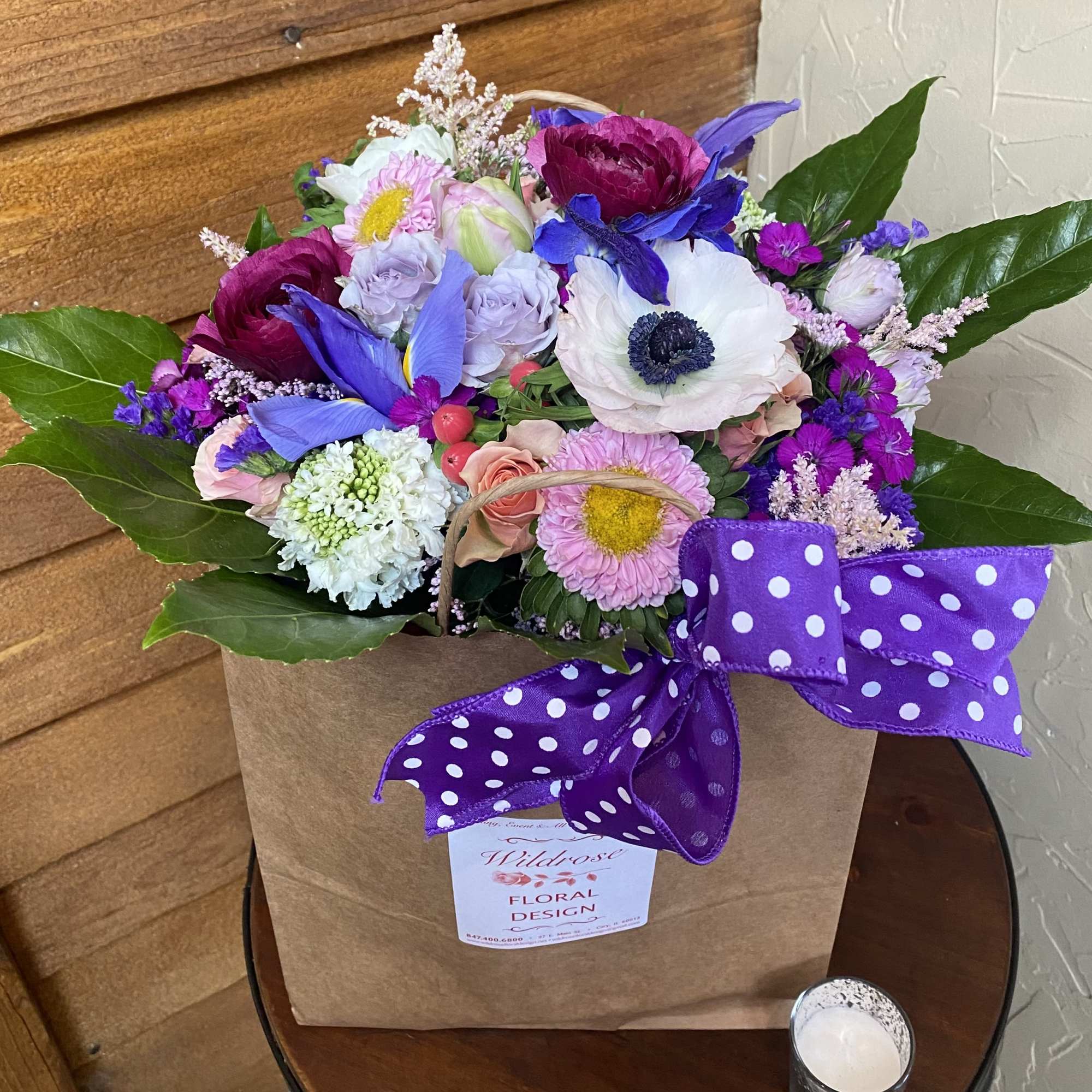 Colorful mixed flower arrangement in a kraft bag with a purple polka dot bow.