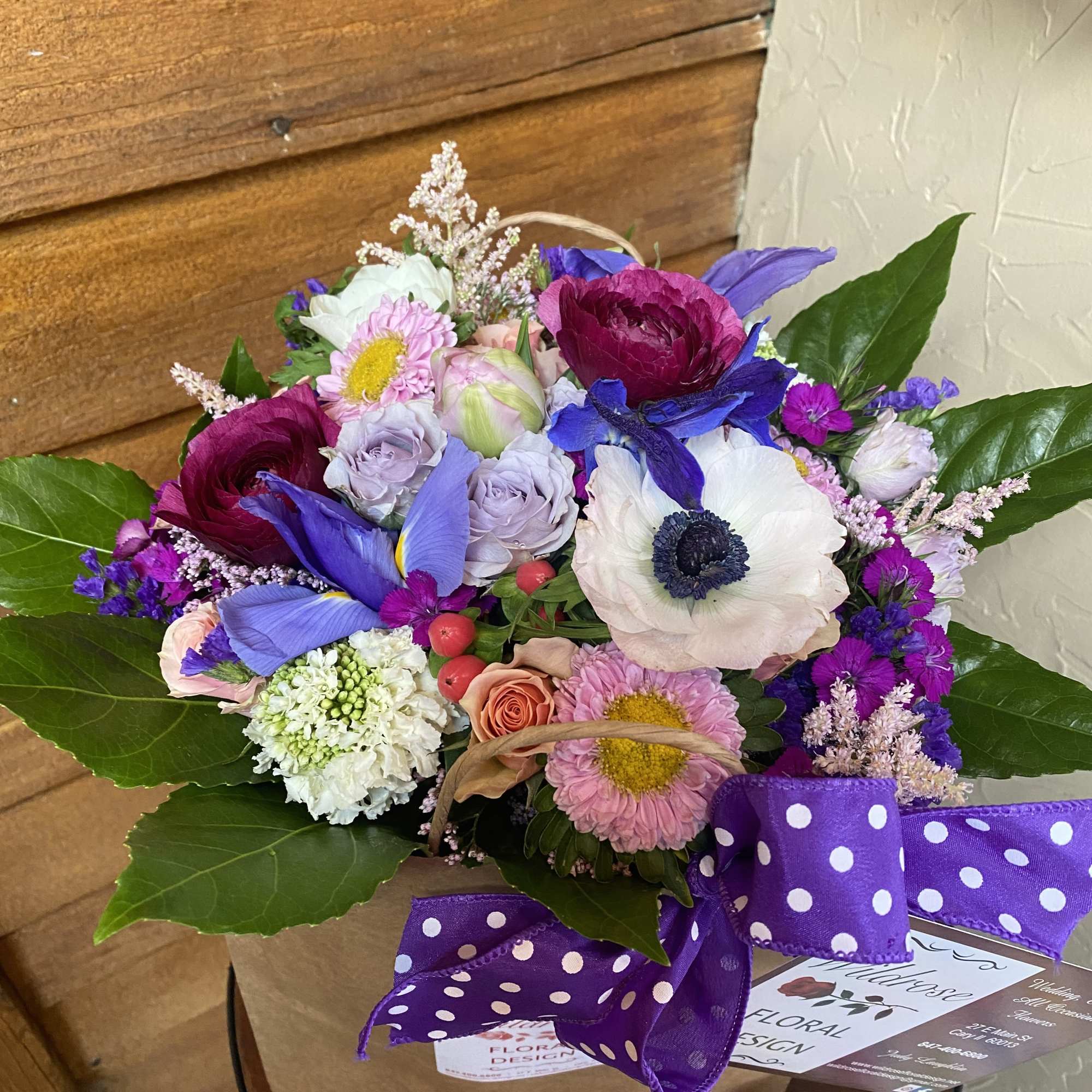 Vibrant mixed bouquet with purple irises, white anemones, and pink blooms in a kraft bag with a polka-dot bow.