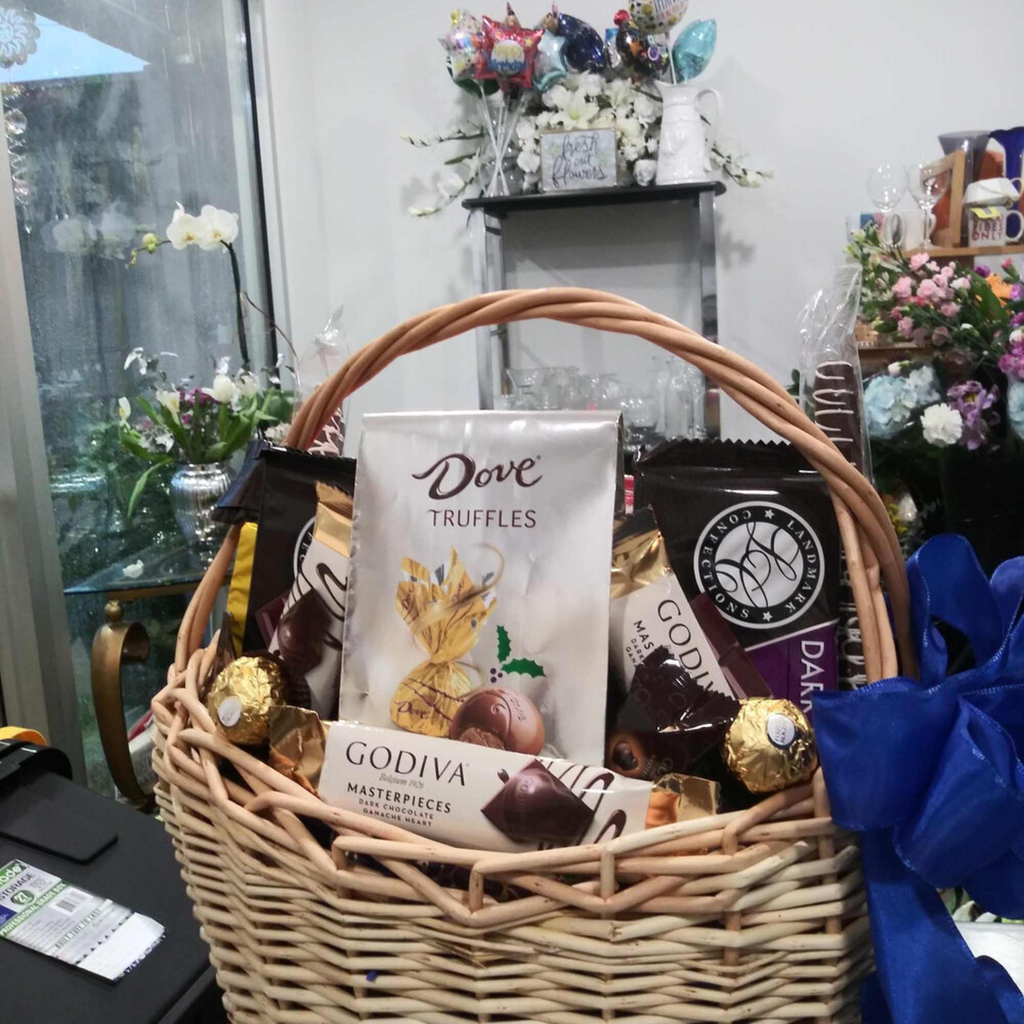 Gift basket with chocolates and a blue ribbon, with flowers in the background