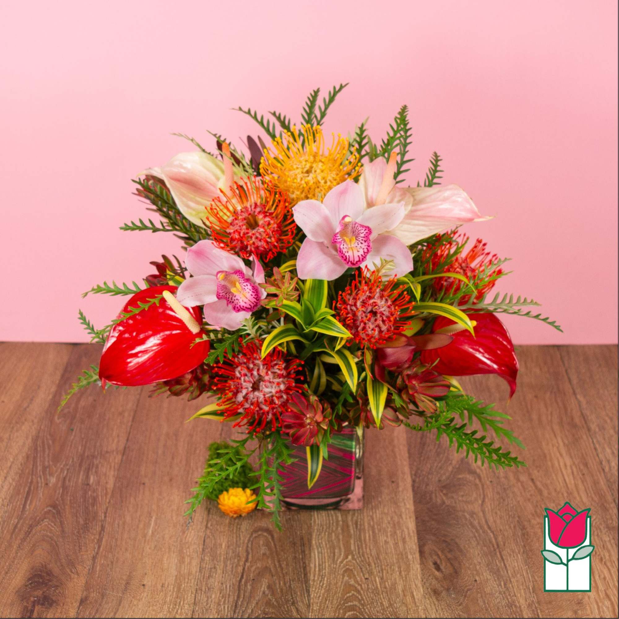 Bright tropical arrangement with red, orange, and pink blooms in a clear glass cube vase on a wooden surface.