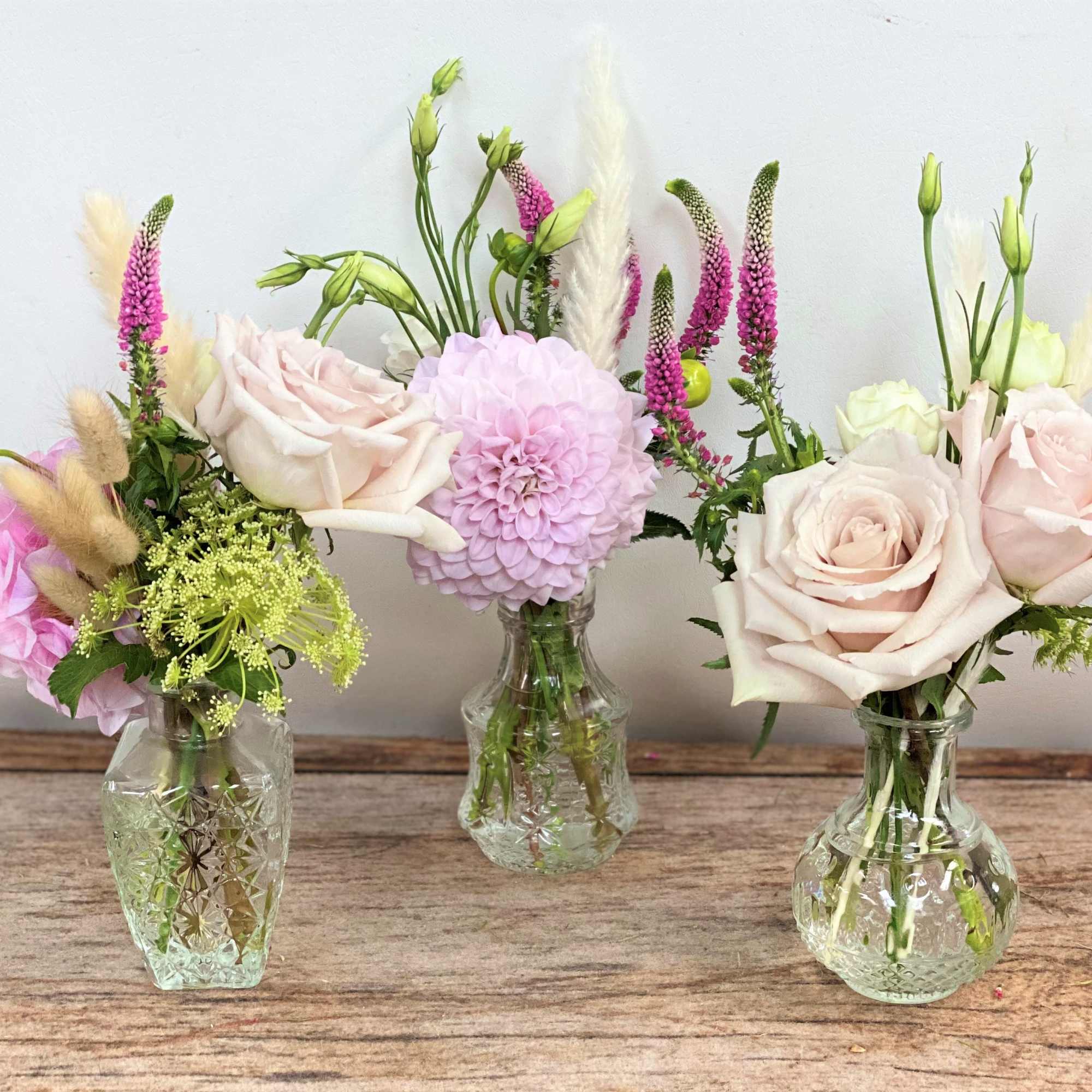 Trio of small glass bud vases with blush roses, a pink dahlia, and delicate accent flowers.