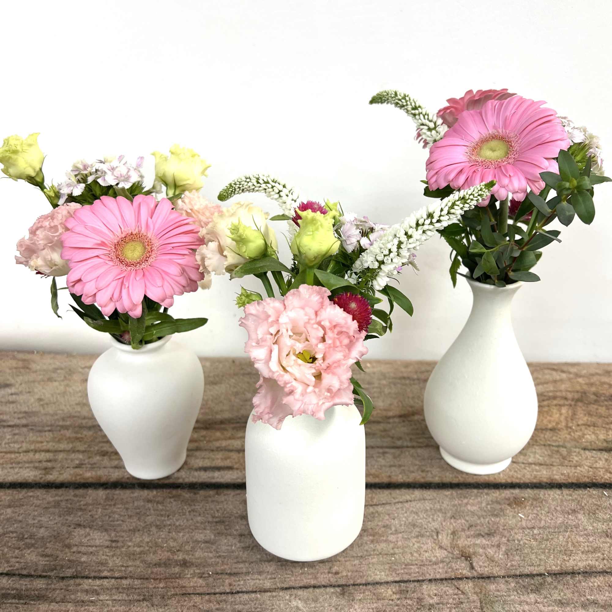 Trio of small white bud vases with pink gerbera daisies and mixed pastel blooms on a wooden surface