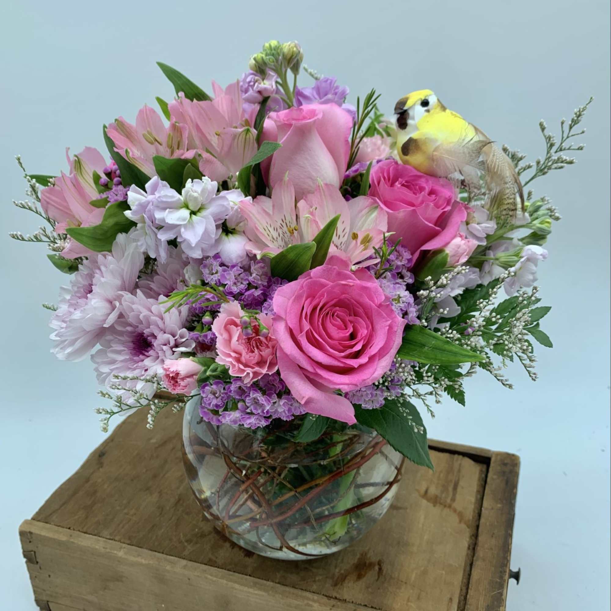 Round glass vase of pink and lavender flowers with a small yellow bird pick on a wooden box