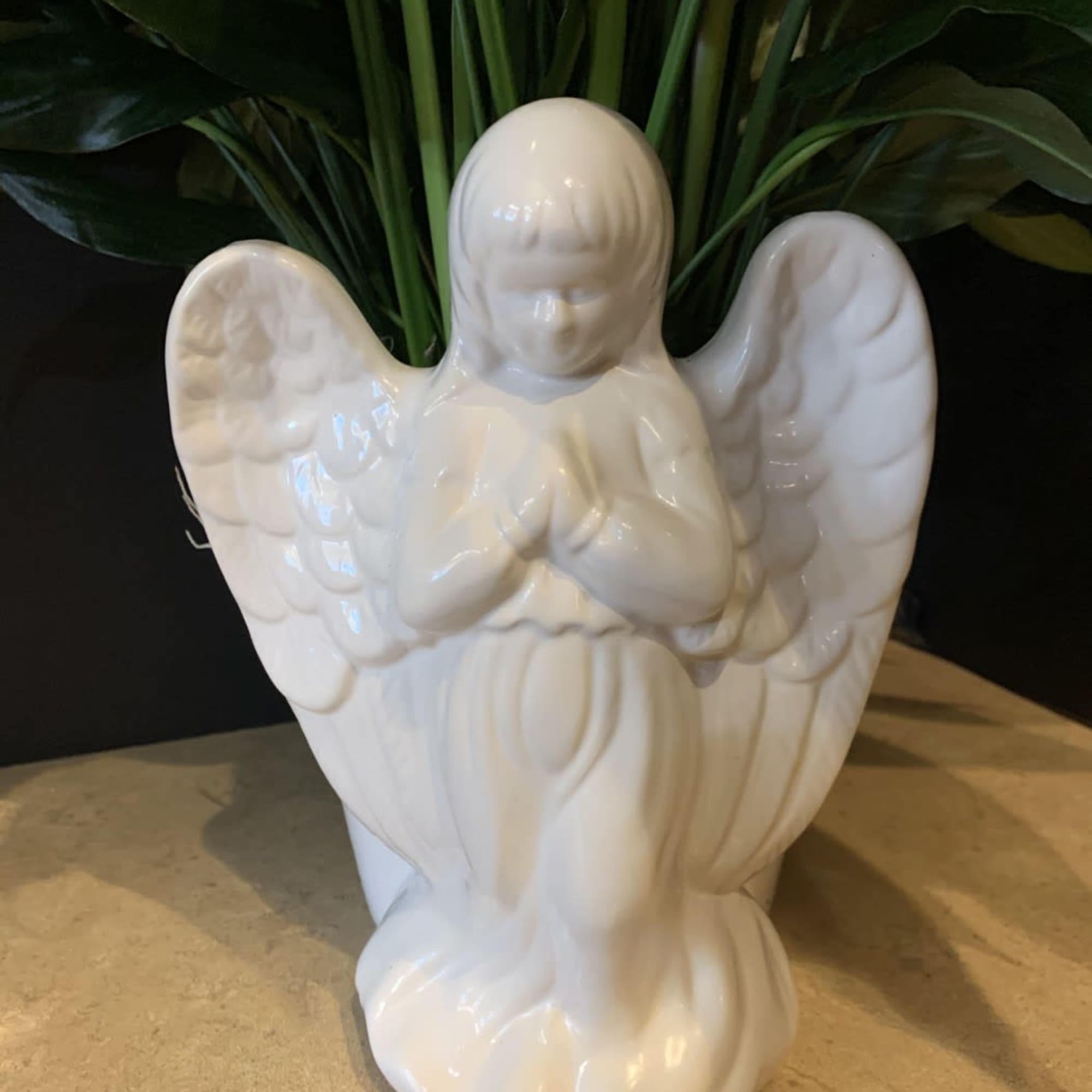 White ceramic angel figurine with folded hands in front of dark green foliage