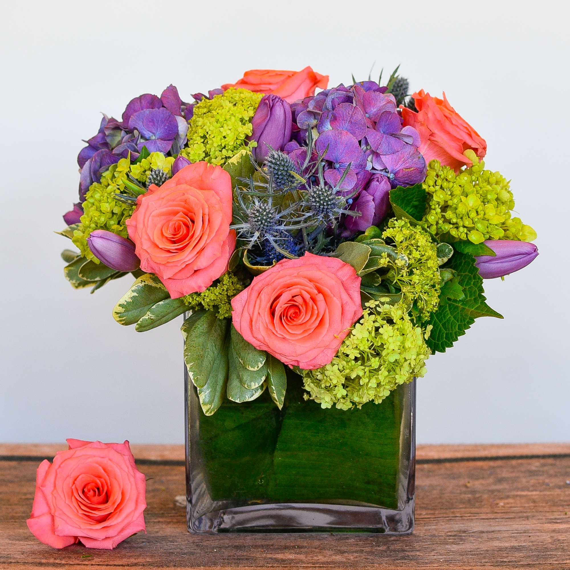 Short arrangement of coral roses, purple blooms, and green hydrangea in a square glass vase with a leaf wrap