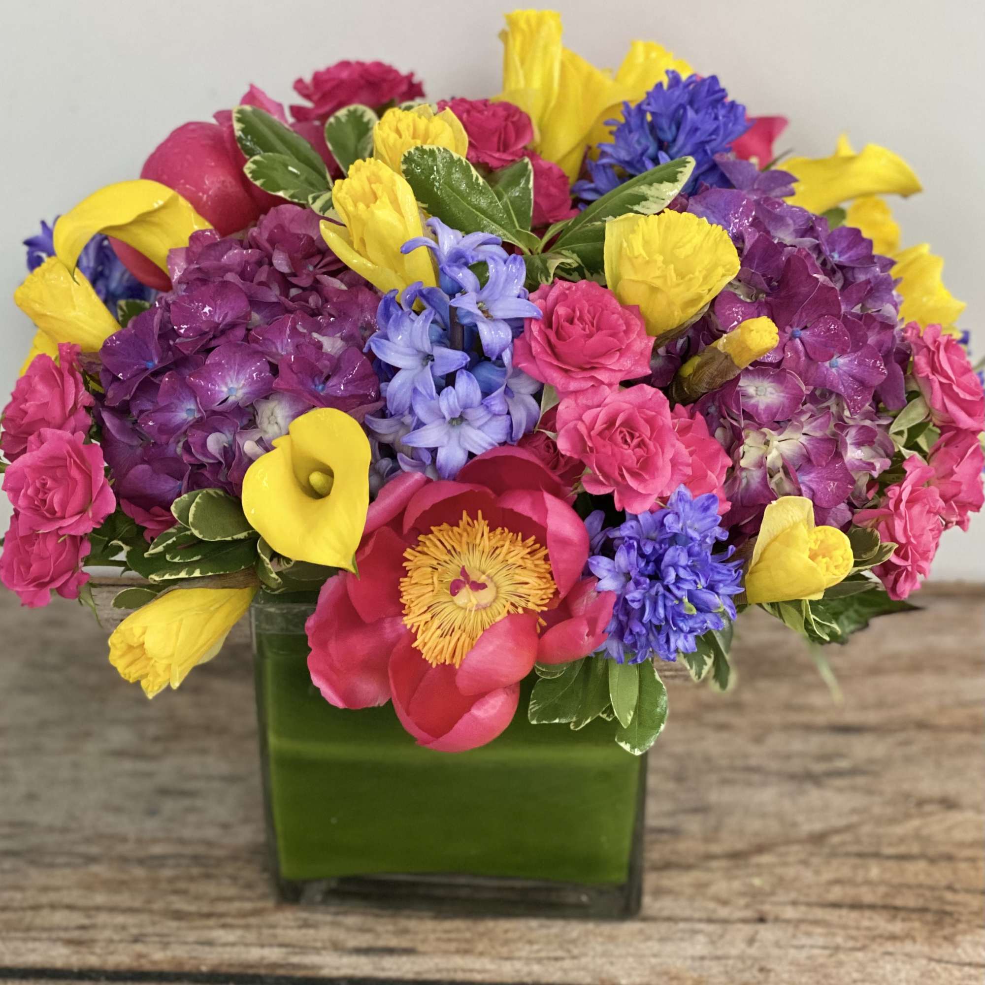 Bright mixed arrangement of pink, yellow, purple, and blue flowers in a square glass vase