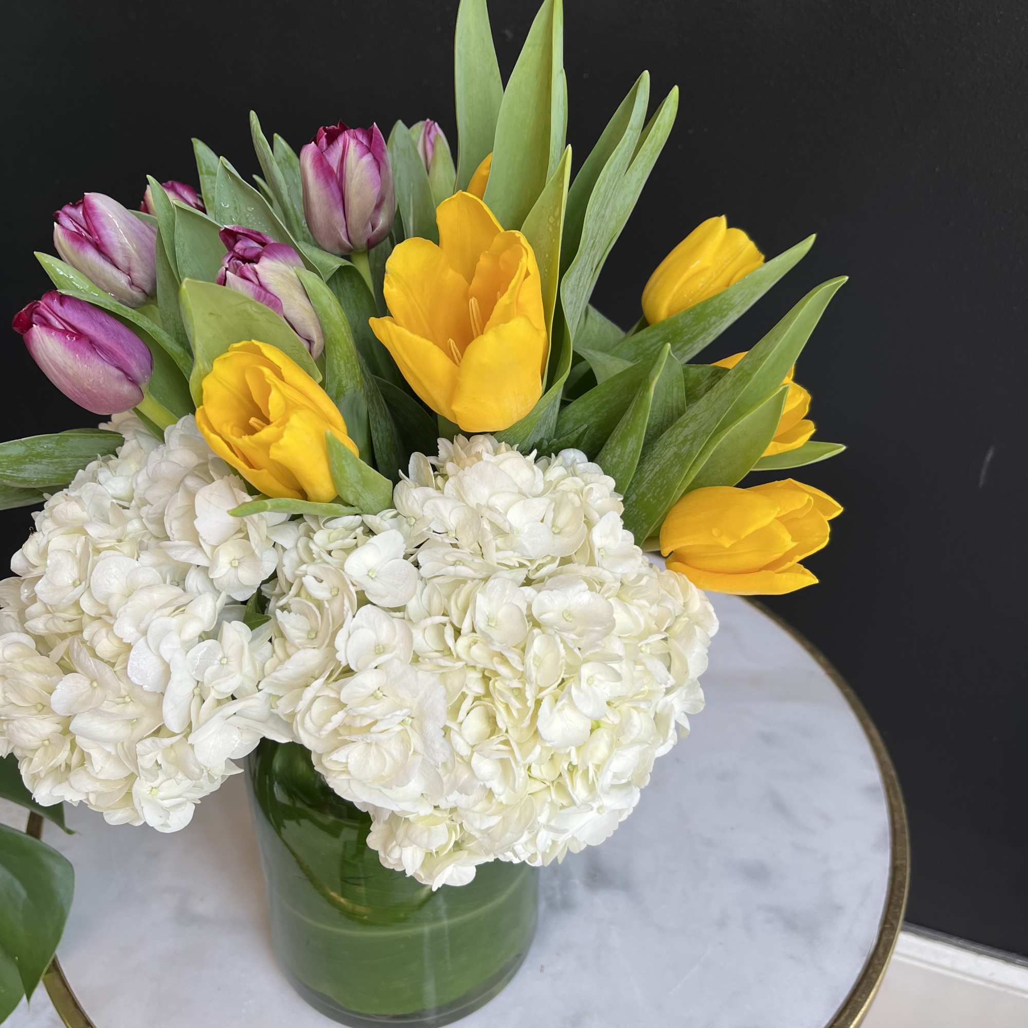 Yellow and purple tulips with white hydrangeas in a green glass vase