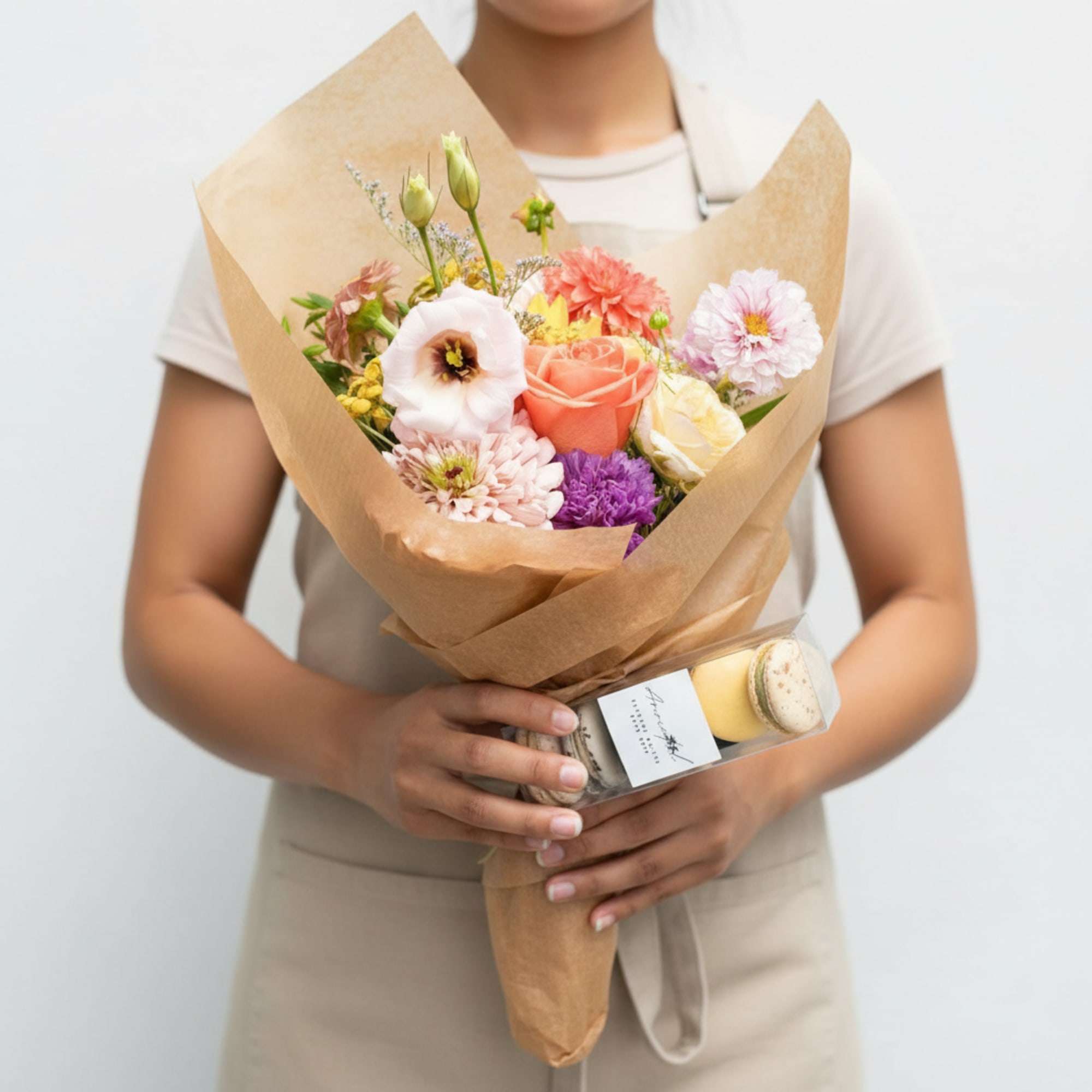 Wrapped pastel flower bouquet with roses and lisianthus held by a person, with a small macaron gift box attached.