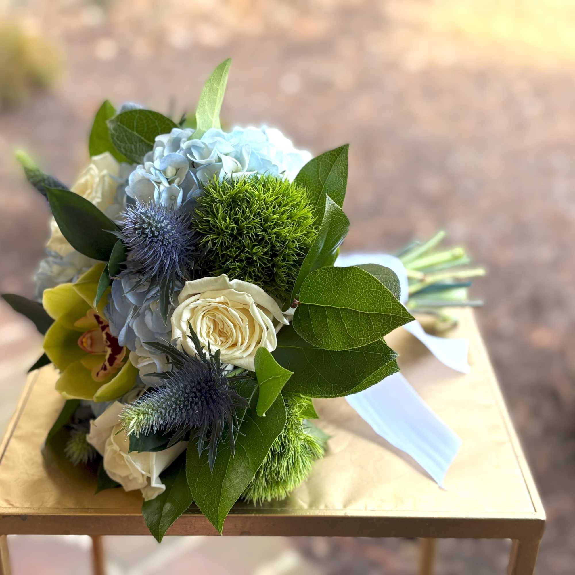 Hand-tied bouquet with blue hydrangea, cream roses, green orchid, and blue thistle on a gold table.