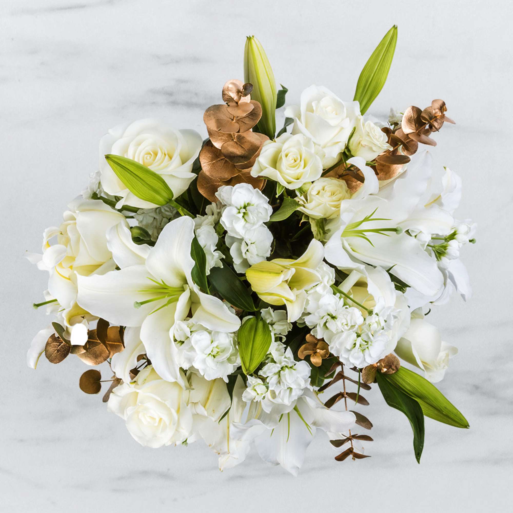 Top view of white lilies, roses, and white stock with metallic gold accents in a compact arrangement.