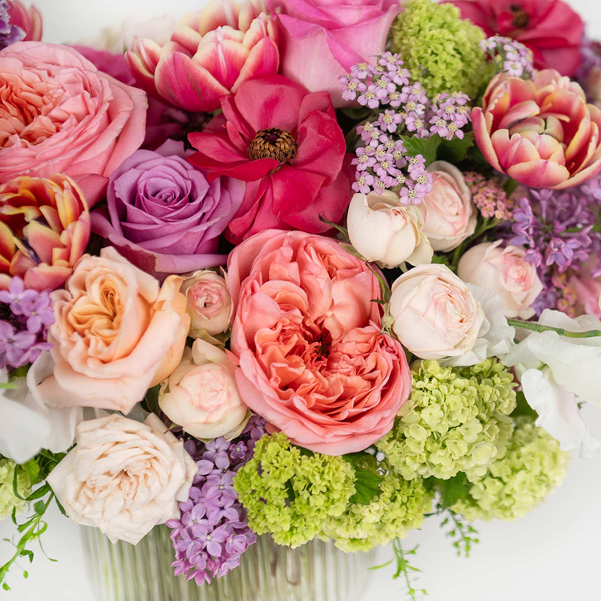 Low arrangement of pink and peach roses with lavender blooms and green hydrangea in a clear glass vase