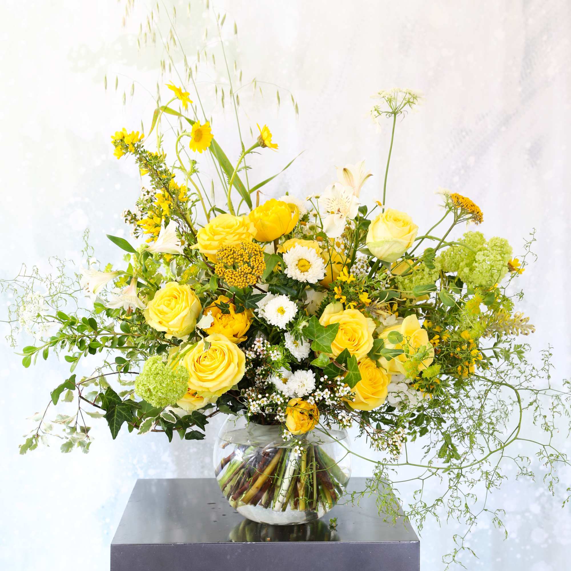 Large yellow and white flower arrangement in a round glass vase on a dark pedestal