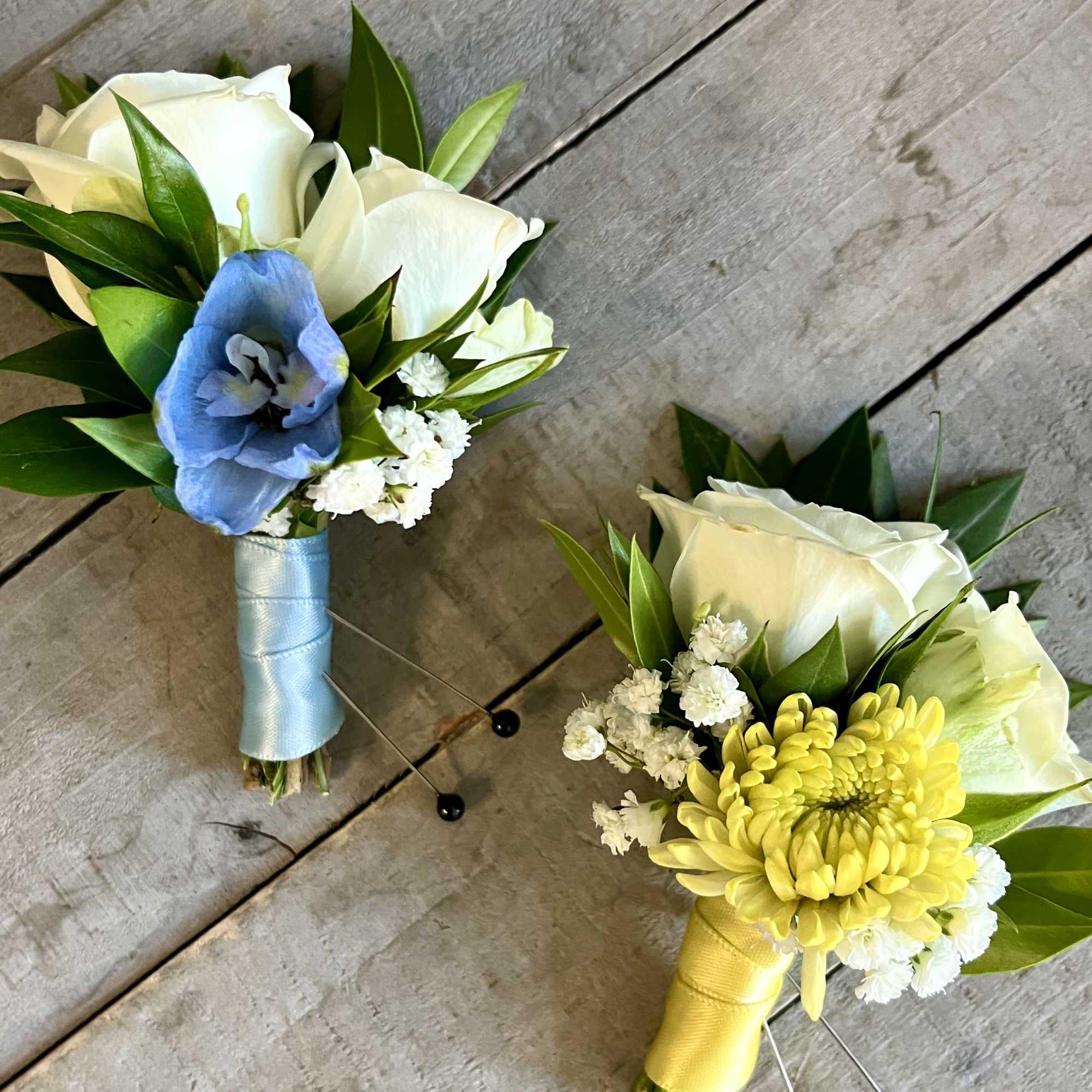 Two small boutonnieres with white roses, one with a blue flower and one with a yellow chrysanthemum.
