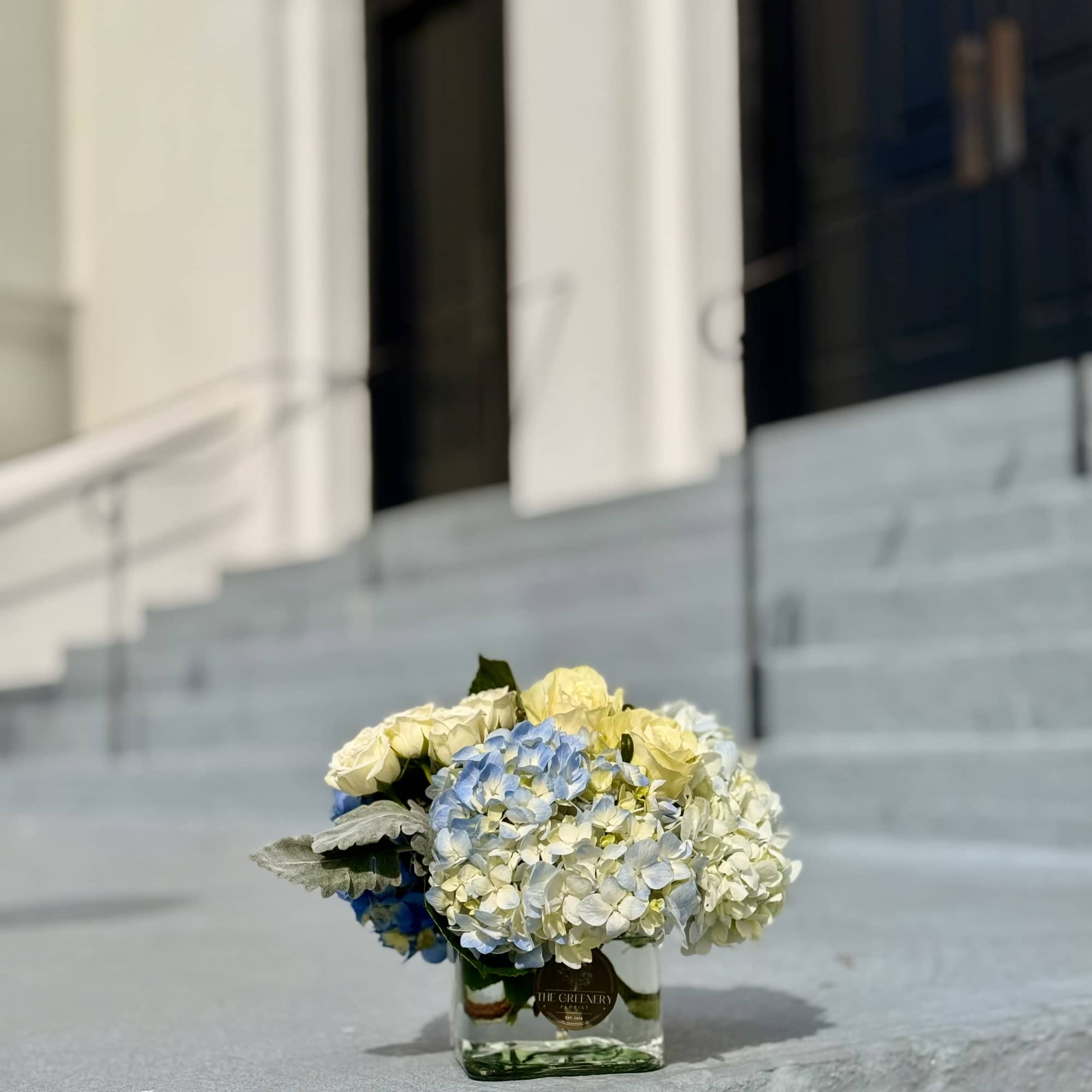 Low arrangement of pale yellow roses and blue and white hydrangeas in a clear square vase on stone steps