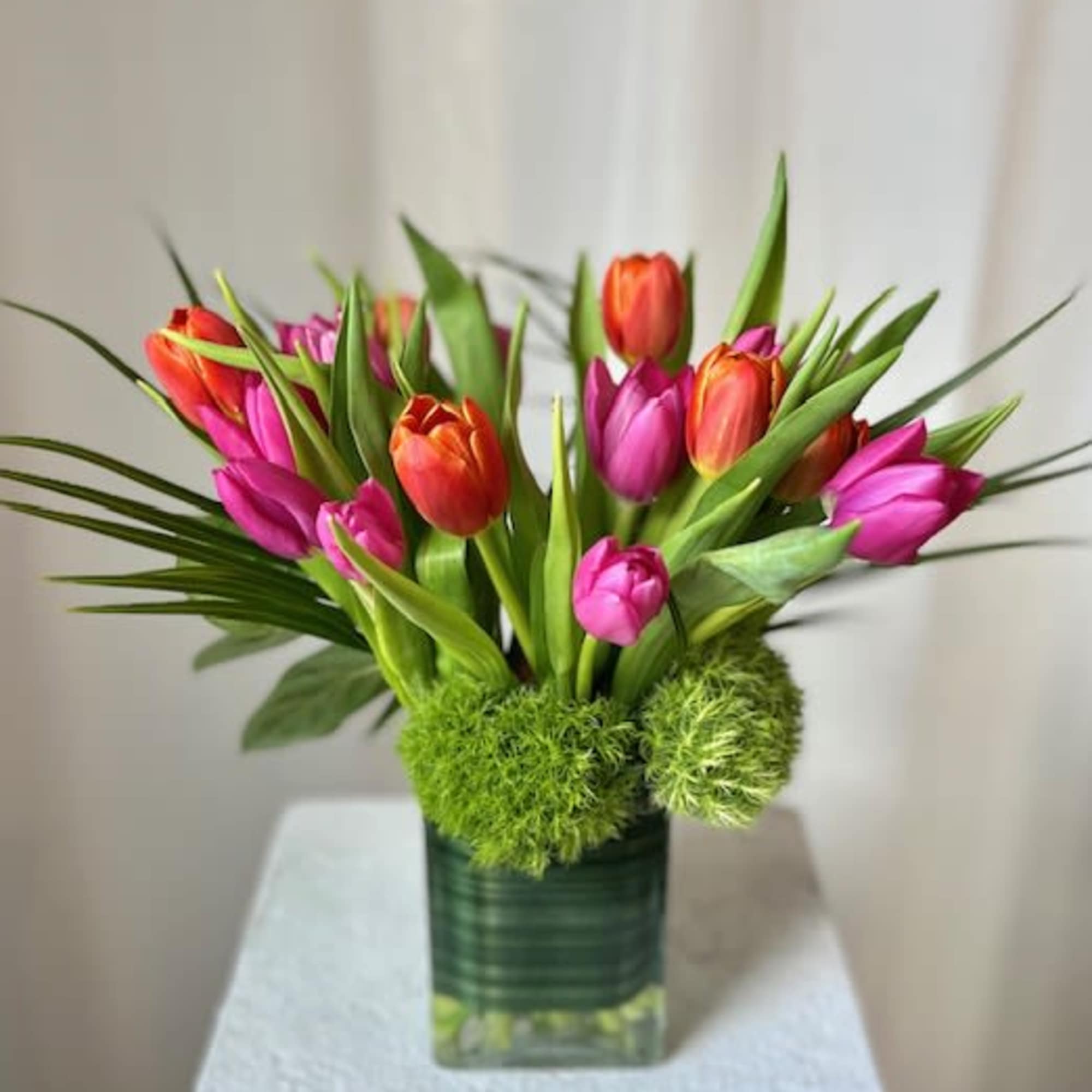 Pink and orange tulips arranged in a square glass vase with green accents