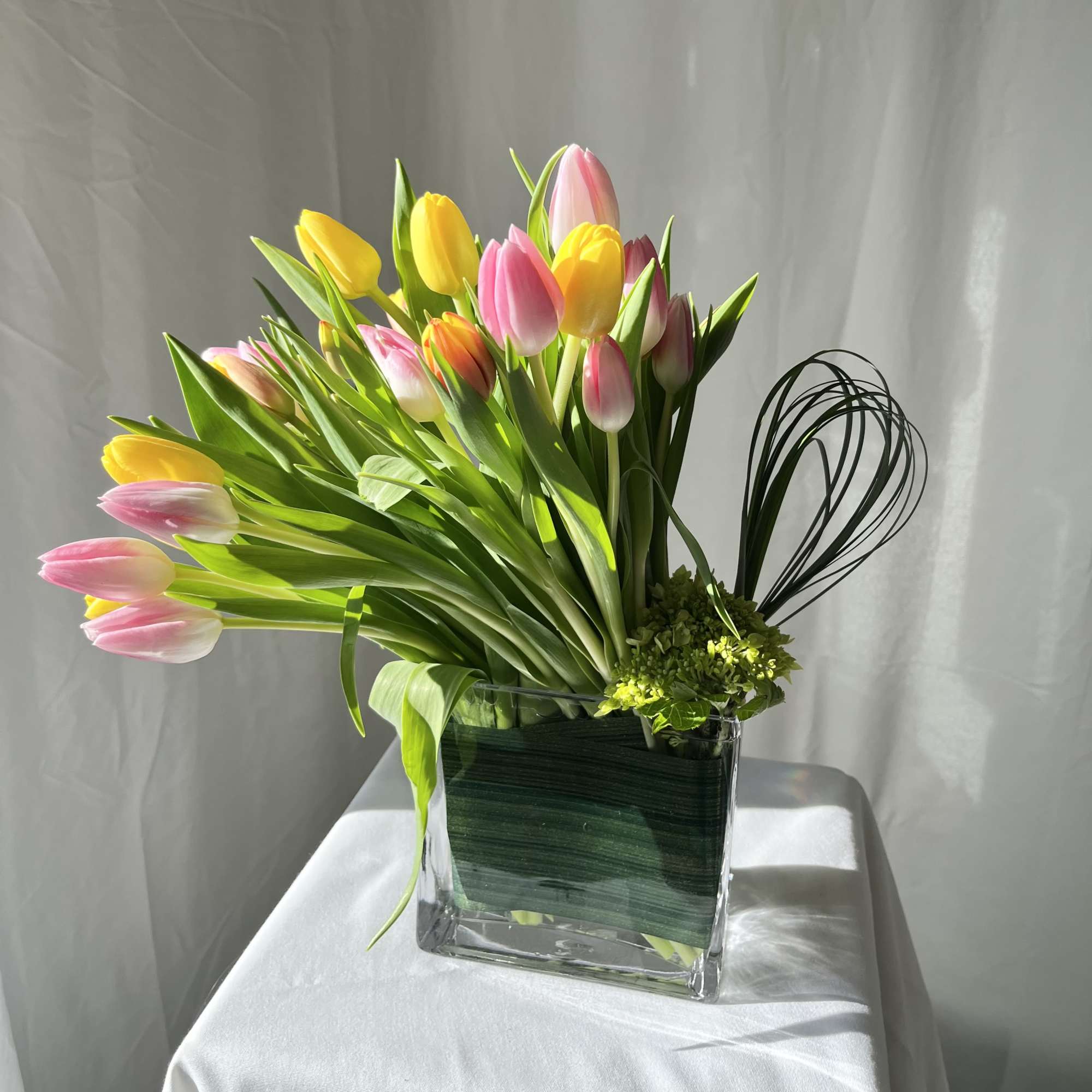 Arrangement of pink and yellow tulips in a clear rectangular glass vase with looped decorative grasses.