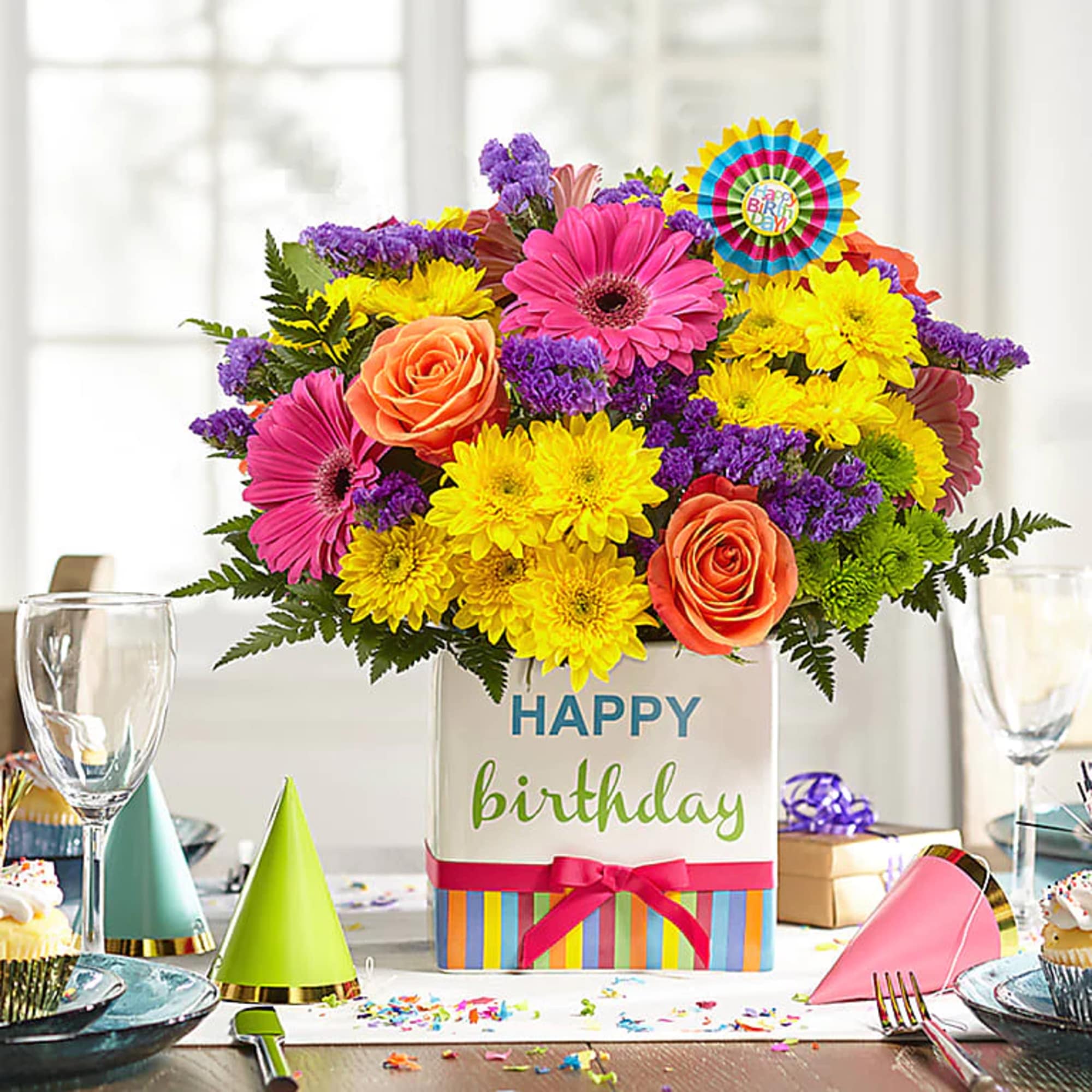 Bright birthday flower arrangement with pink gerberas, yellow mums, and orange roses in a cube vase.