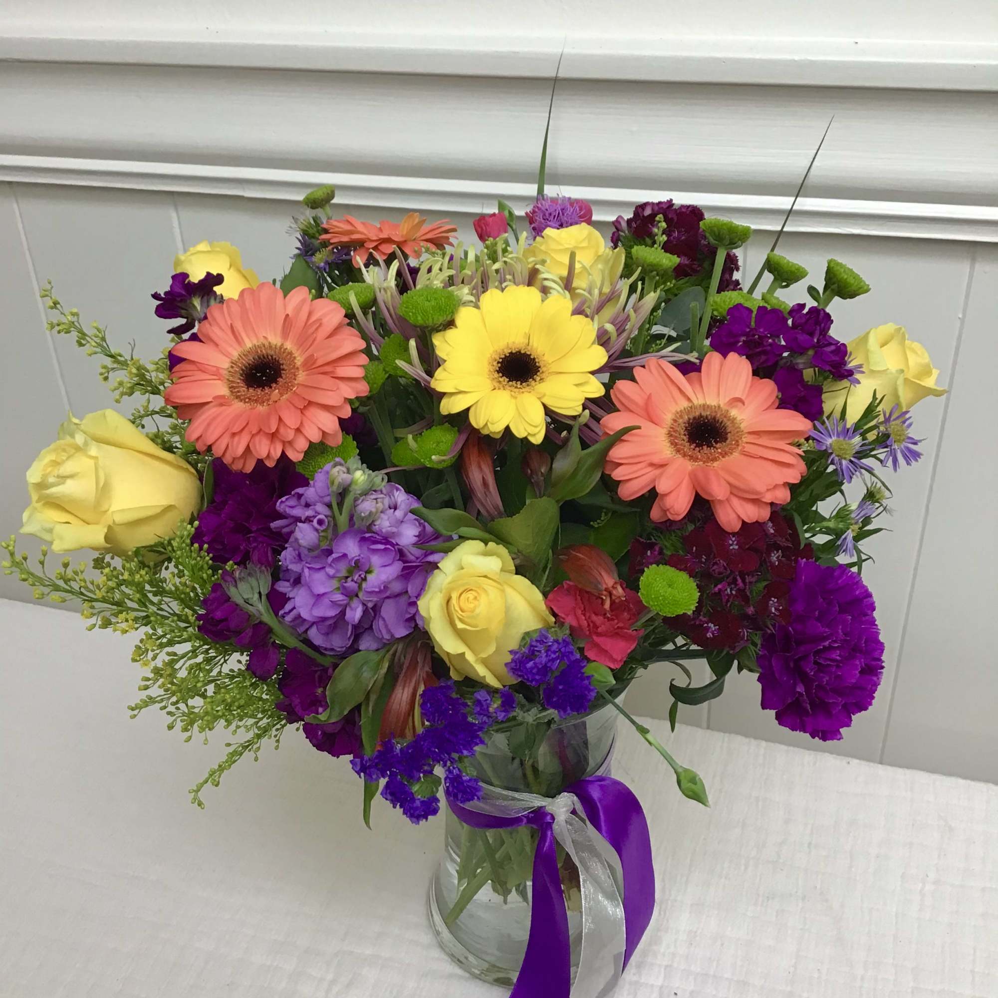 Mixed bouquet of yellow roses, gerbera daisies, and purple flowers in a glass vase with a ribbon.