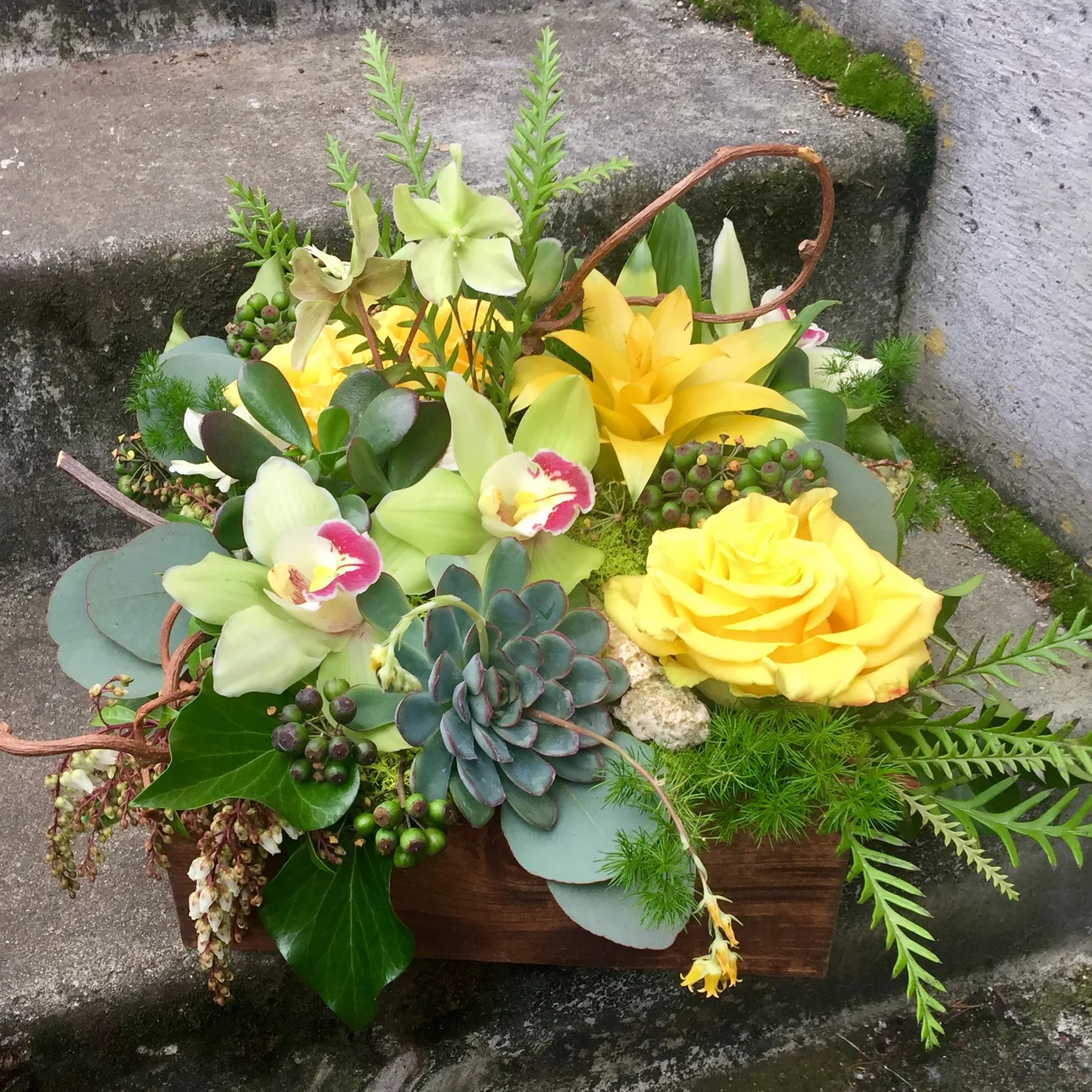Low wooden box arrangement with yellow roses, green orchids, and succulents.