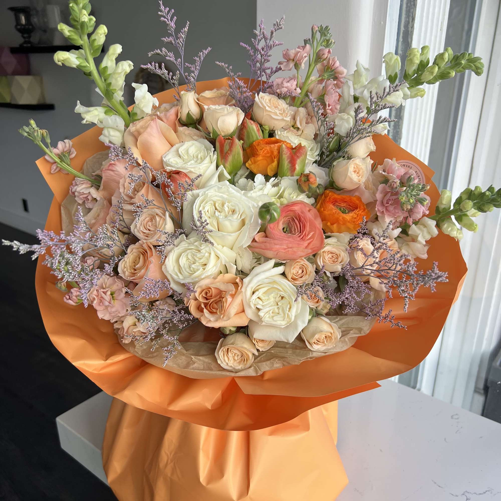 Hand-tied bouquet of peach and white roses with orange ranunculus wrapped in bright orange paper.