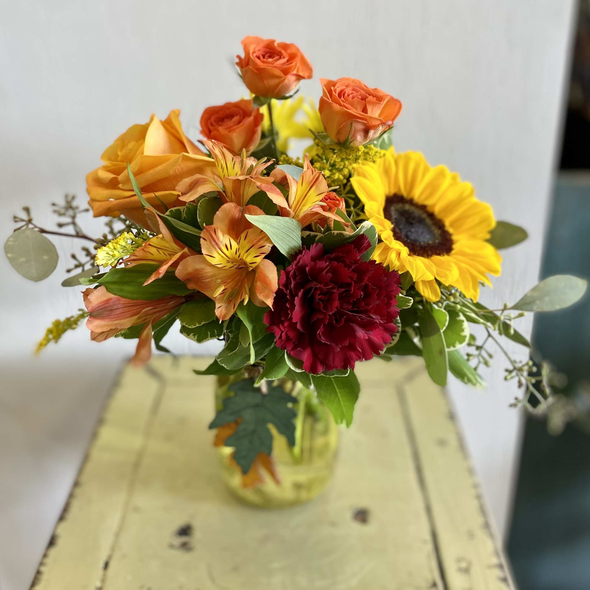 Mixed bouquet with orange roses, sunflowers, and red carnations in a glass vase