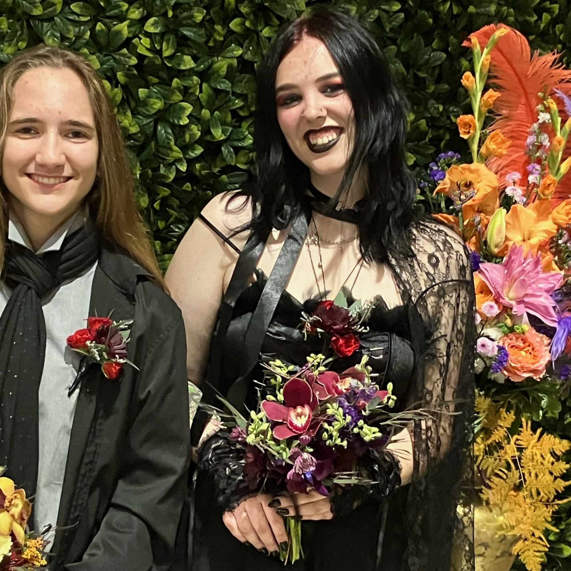 Two people in dark formal outfits holding floral bouquets in front of a leafy wall.